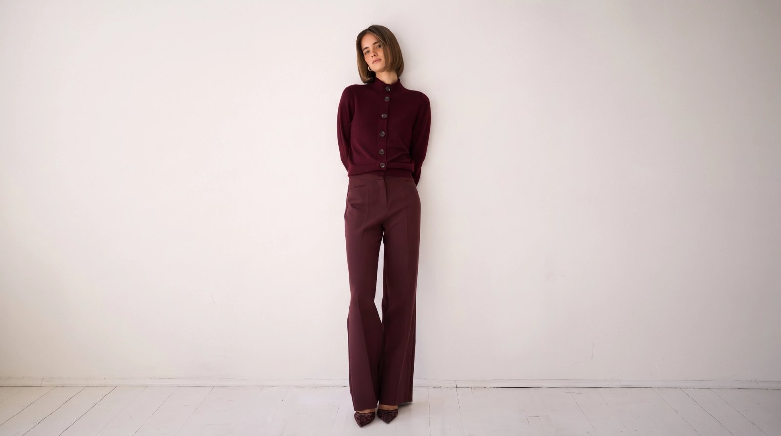 A woman wearing a burgundy cardigan and flared pants that match stands against a white wall.