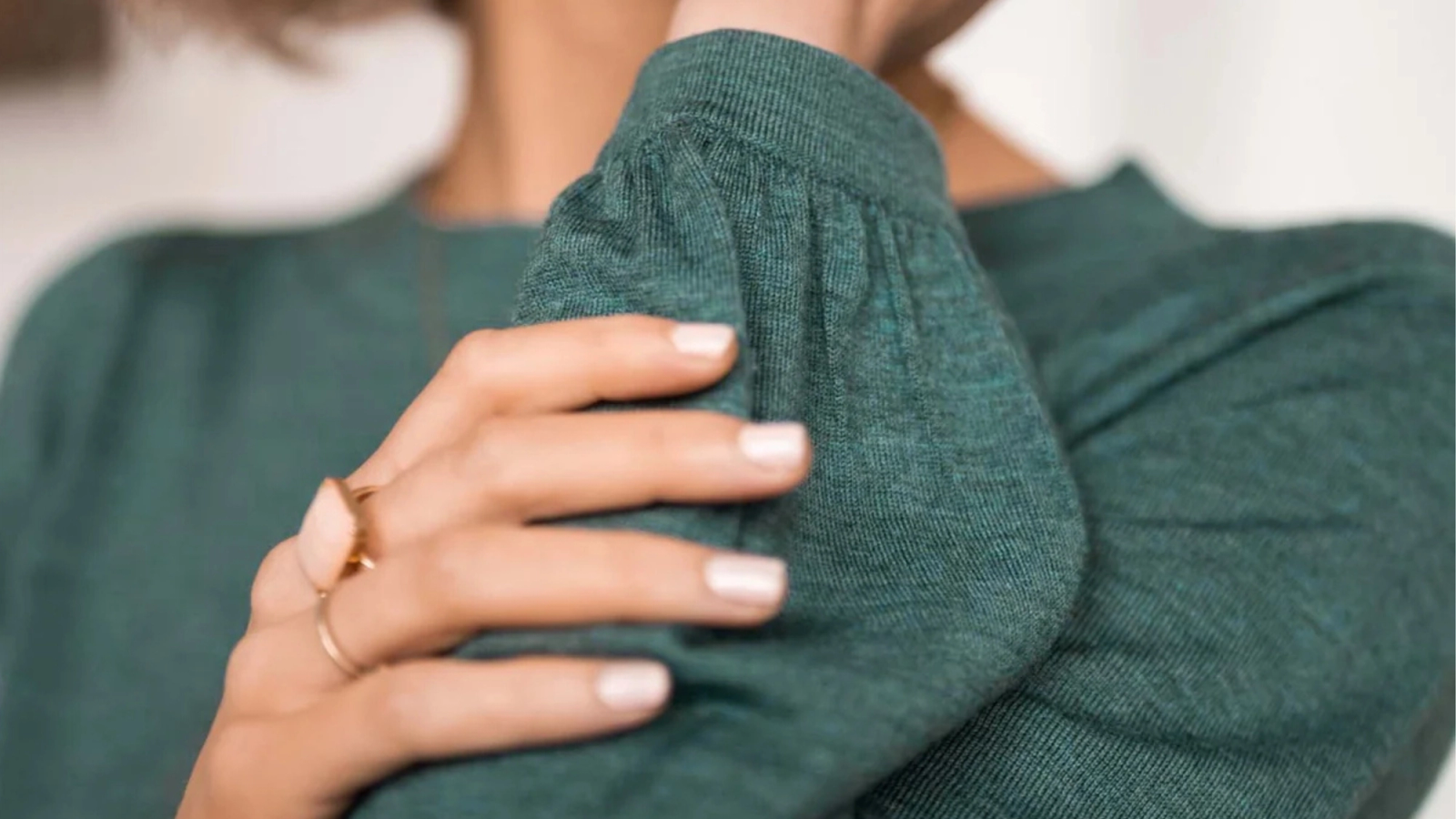 A hand with gold rings and light-colored nail polish resting on a dark green knit top with gathered sleeves.