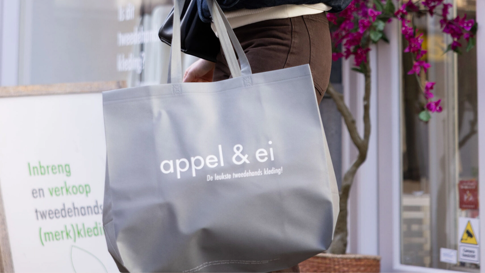 A person holds a grey 'appel & ei' tote bag in front of a second-hand clothing store.