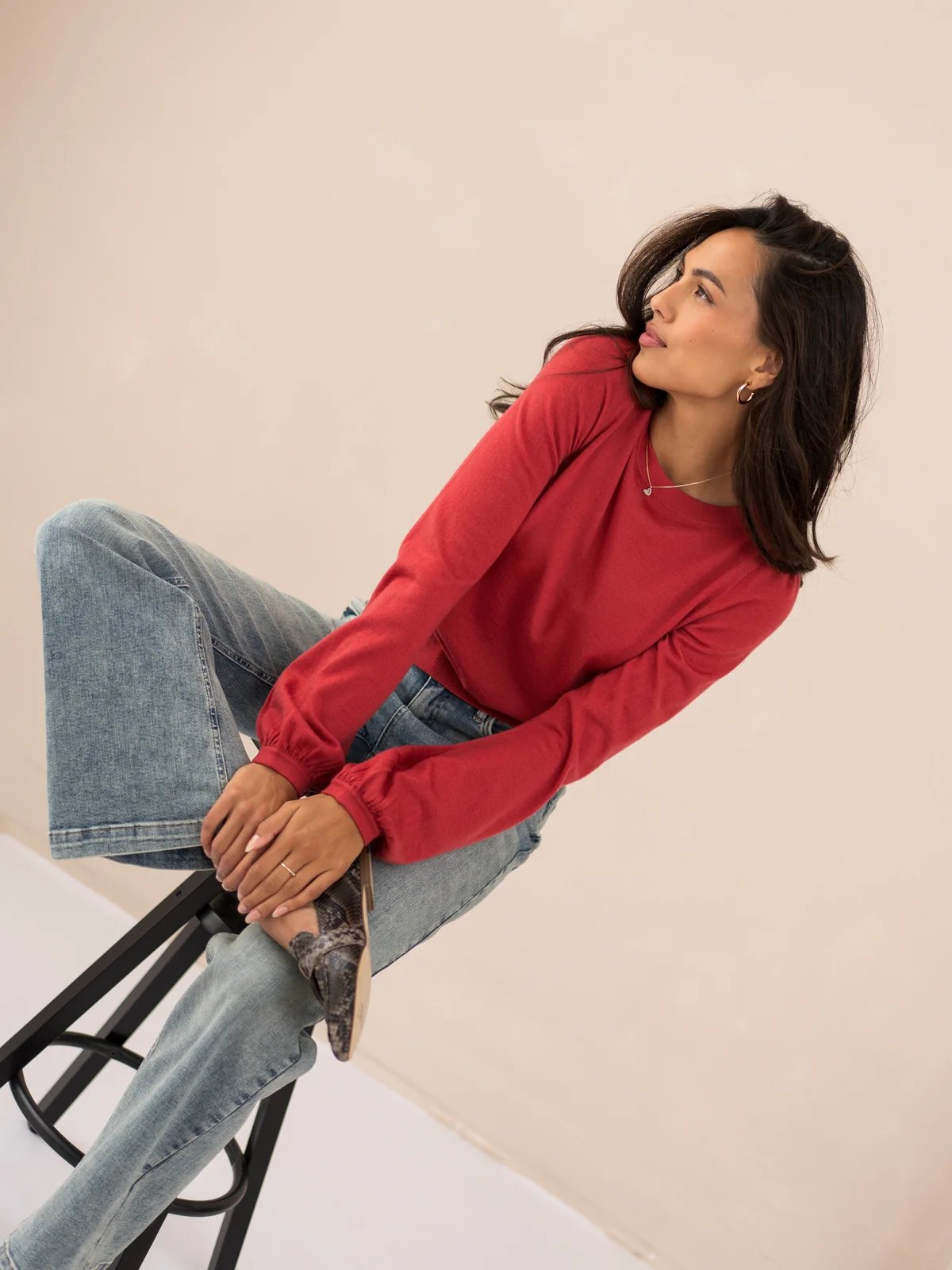 A woman wearing a red long-sleeve top and blue jeans is sitting on a stool, looking to the right.