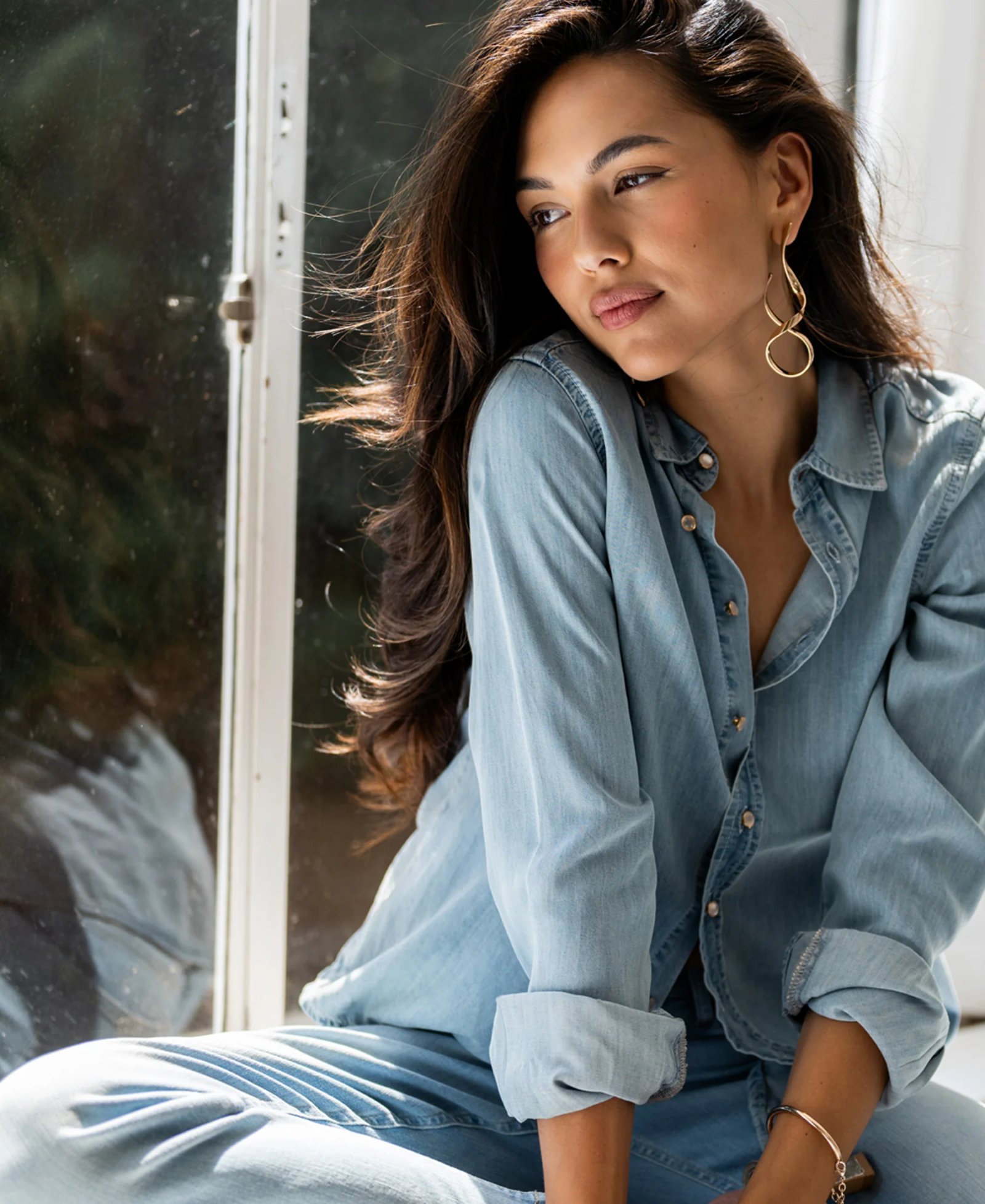 A woman in a light blue denim shirt and jeans, wearing abstract gold dangle earrings, sits thoughtfully by a window.