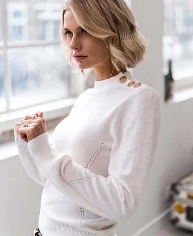 Eine blonde Frau in einem weißen Pullover mit Stehkragen, goldenen Schulterknöpfen und perforierten Details, die nachdenklich nach links blickt.