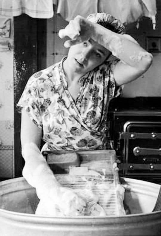 Black-and-white photo of a woman hand-washing laundry with a washboard, wiping suds from her forehead.
