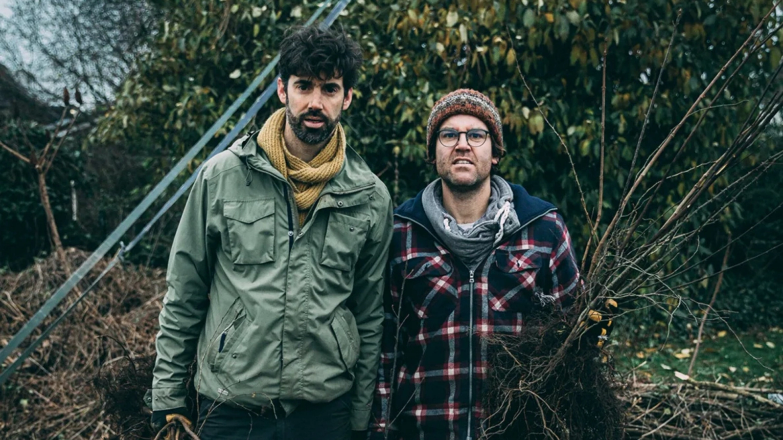 Two men standing outdoors, one holding a bundle of bare-root saplings.