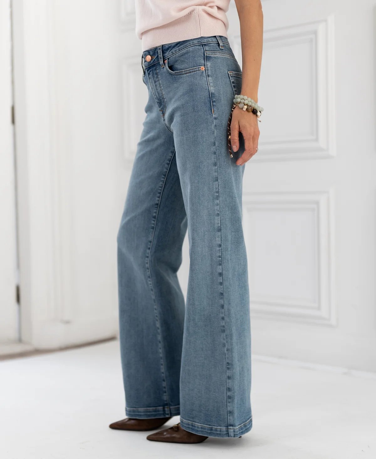 A person stands wearing light blue wide-leg jeans, a pink top, brown shoes, and beaded bracelets.