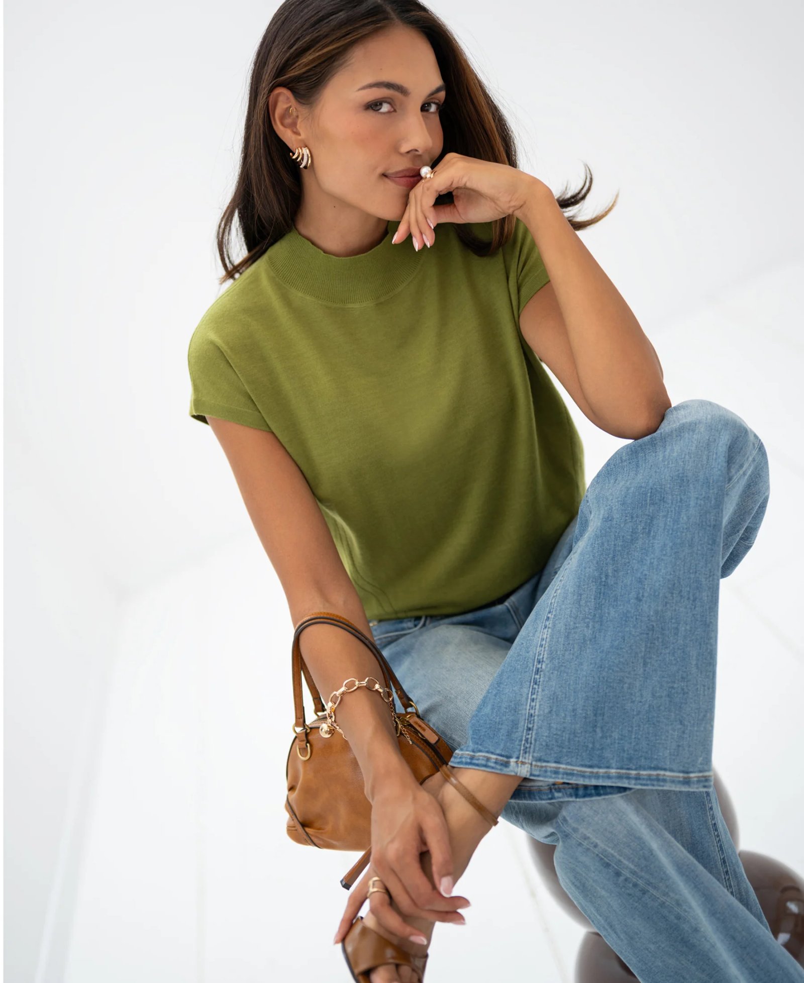 A woman wearing a green mock-neck top, blue jeans, and brown sandals looks at the camera; she has a pearl ring on her finger and is holding a small brown handbag.