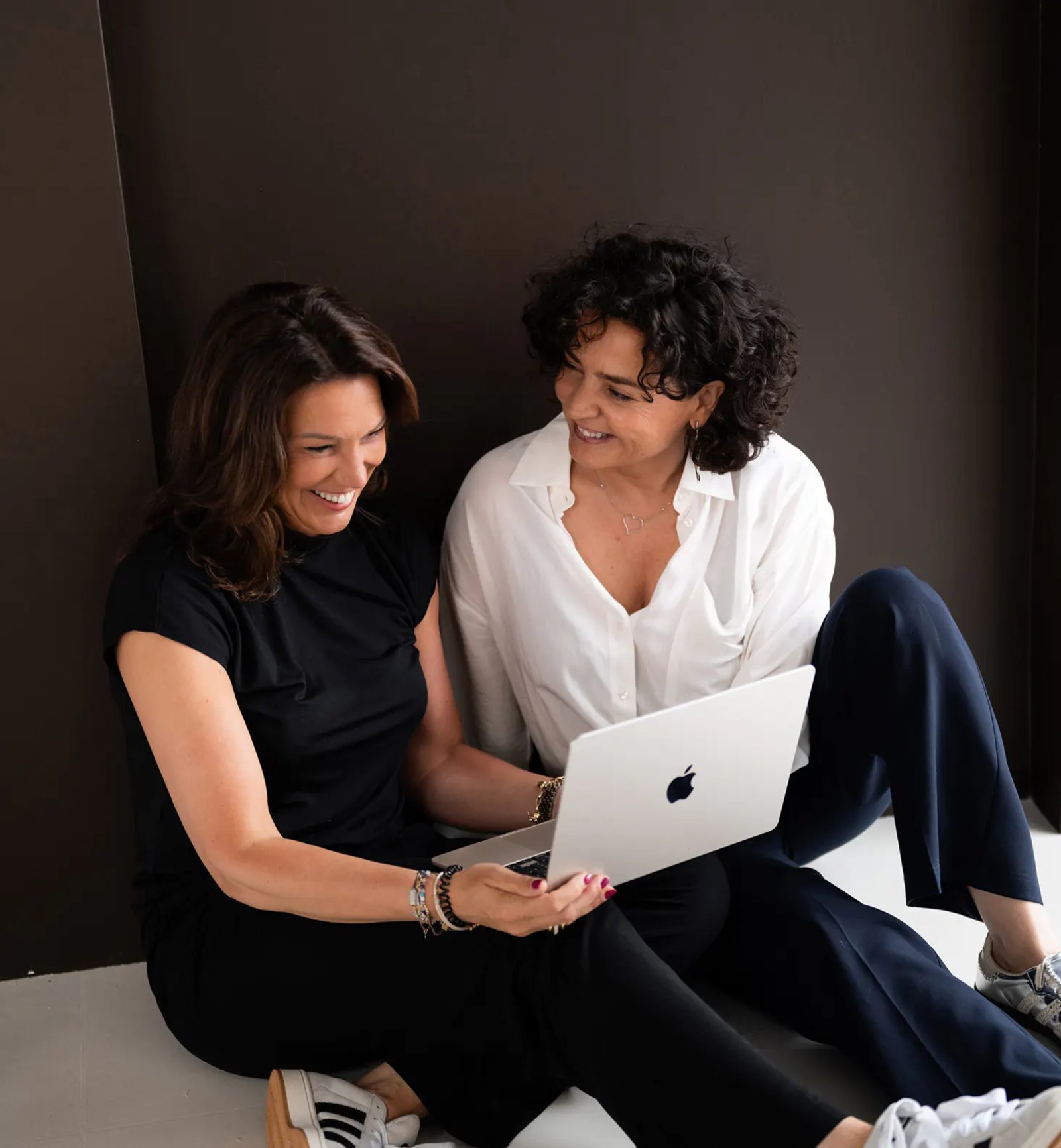 Zwei lächelnde Frauen sitzen auf dem Boden und schauen auf einen Laptop.