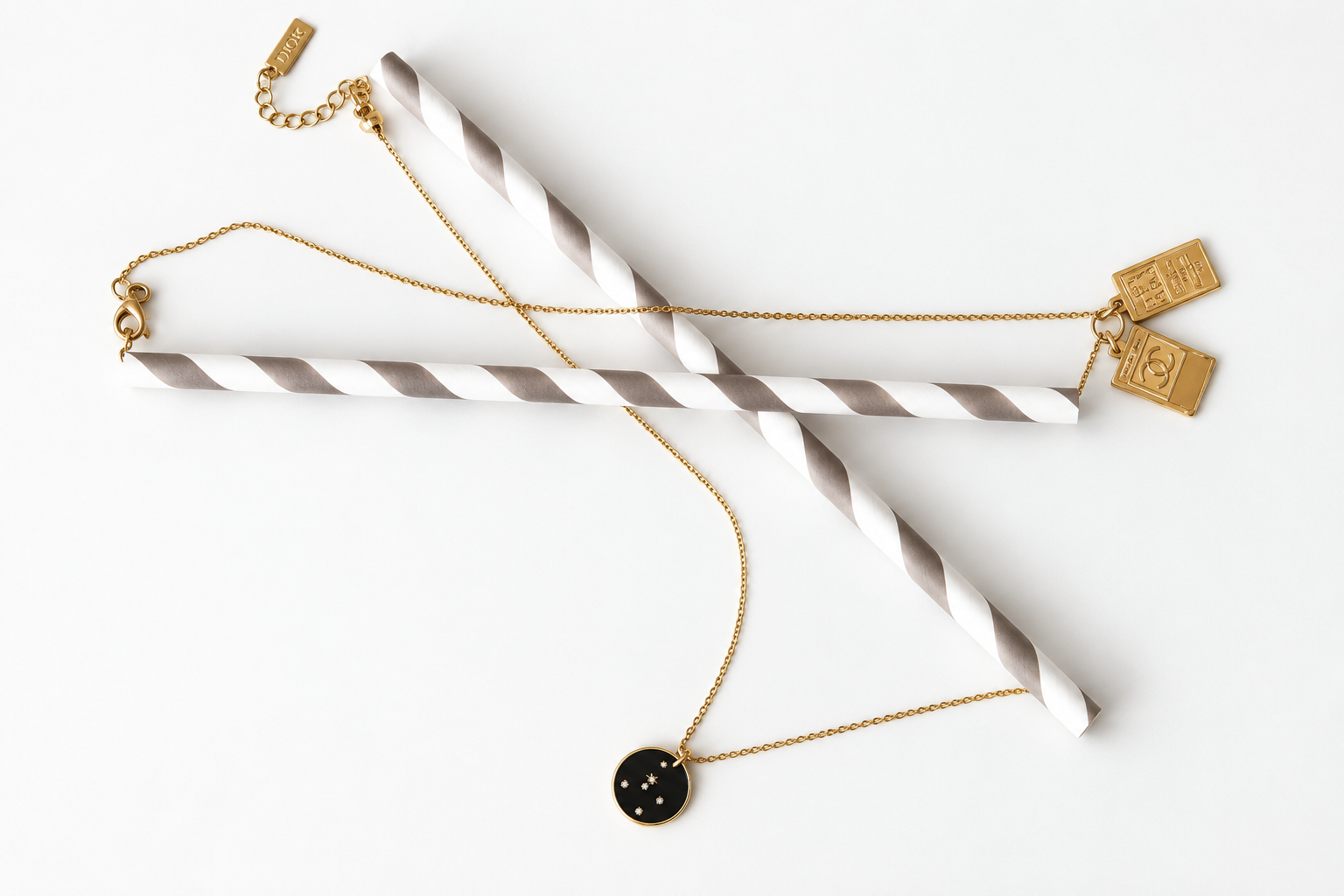 Gold necklaces, one with a Chanel-logo charm and one with a black constellation pendant, draped over three grey and white striped paper straws on a white background.