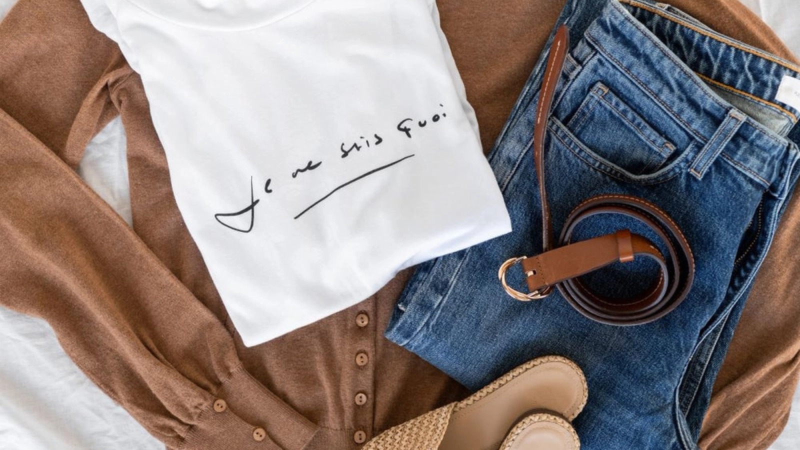 Flatlay einer braunen Strickjacke, eines weißen „Je ne sais quoi“-T-Shirts, einer blauen Jeans, eines braunen Gürtels und gewebter Pantoletten.