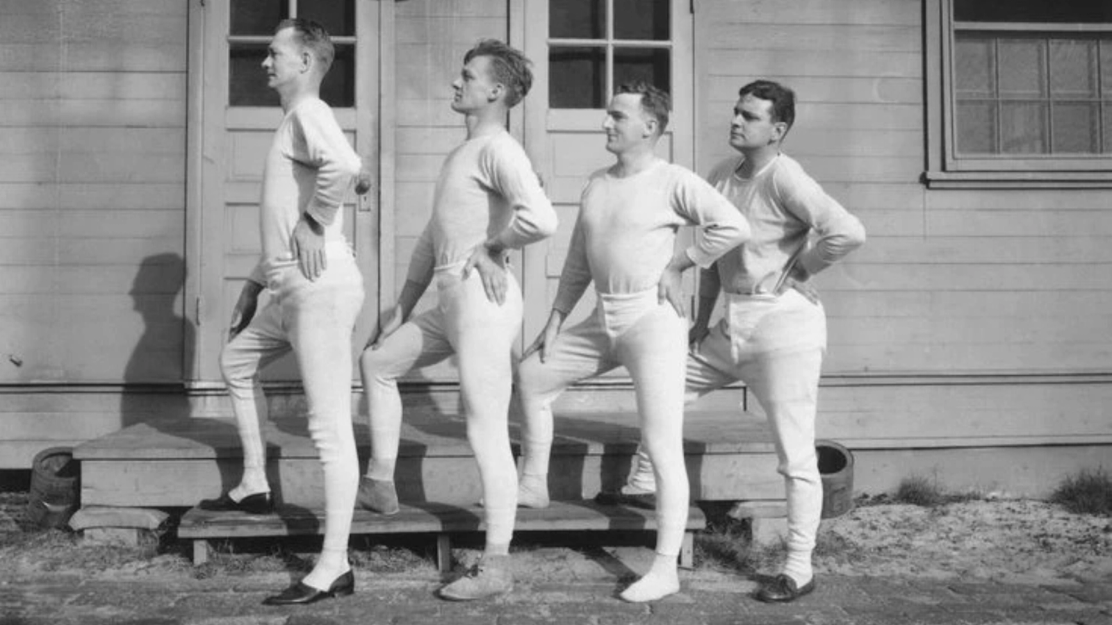 Four men in long johns pose outdoors, striking a theatrical pose with one leg bent.