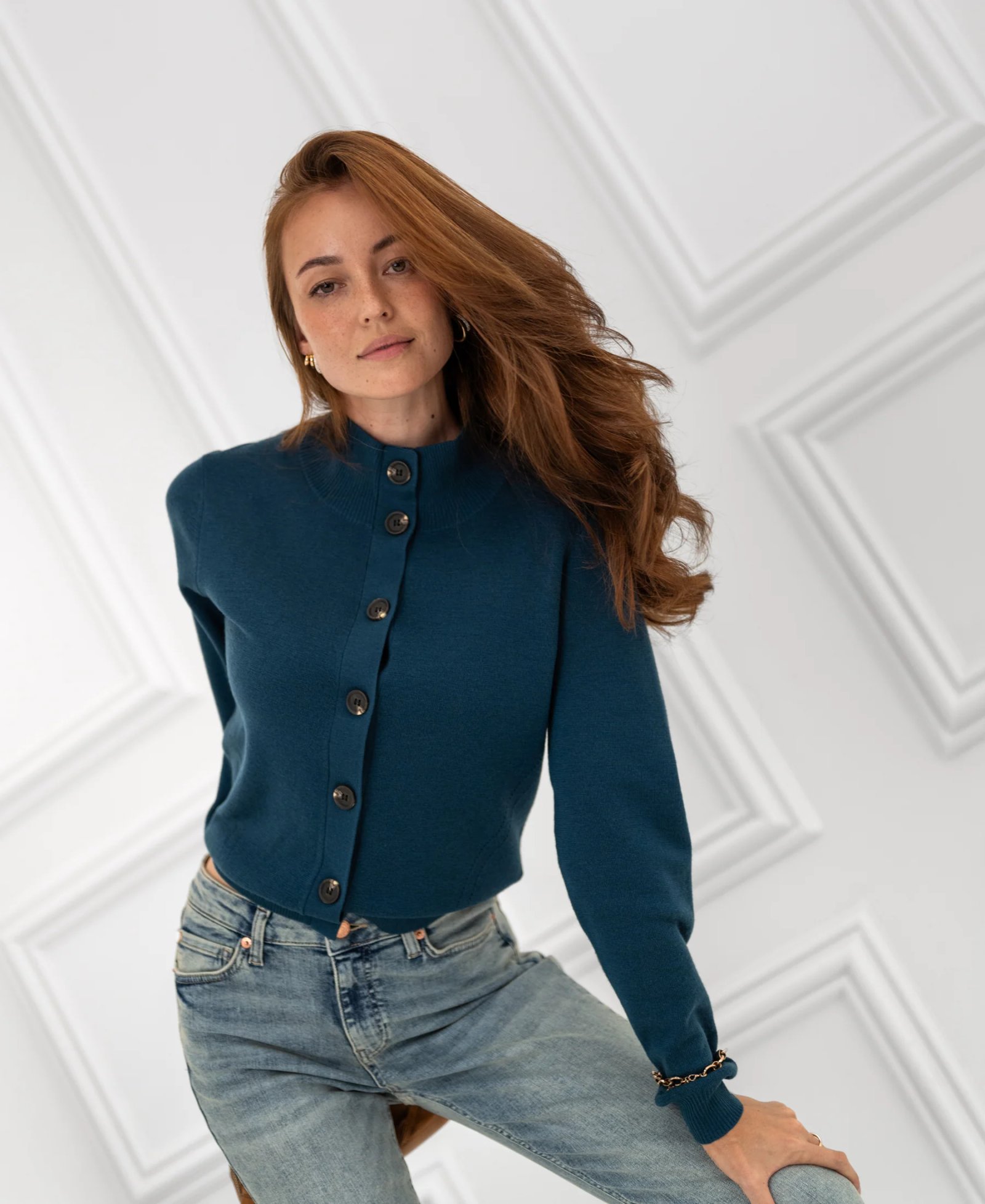 A woman with long red hair wears a teal button-up cardigan and light-wash jeans, looking at the camera.