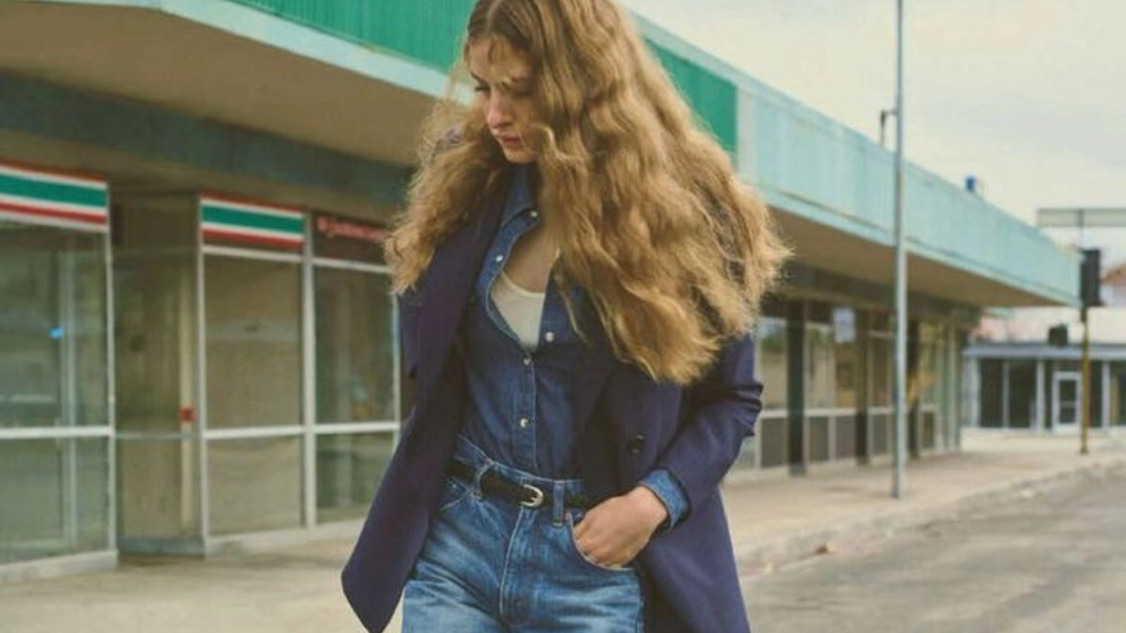 A young woman with long blonde hair, wearing a denim shirt, jeans, and a dark blue coat, walks past a 7-Eleven.
