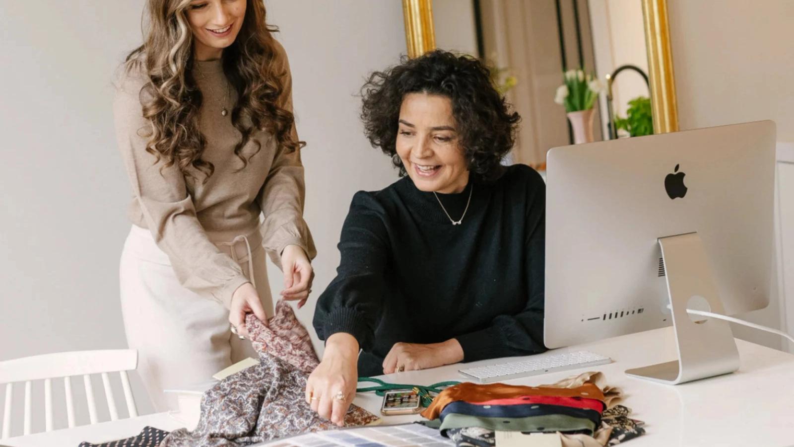 Two smiling women examining fabric swatches at a desk with a computer.
