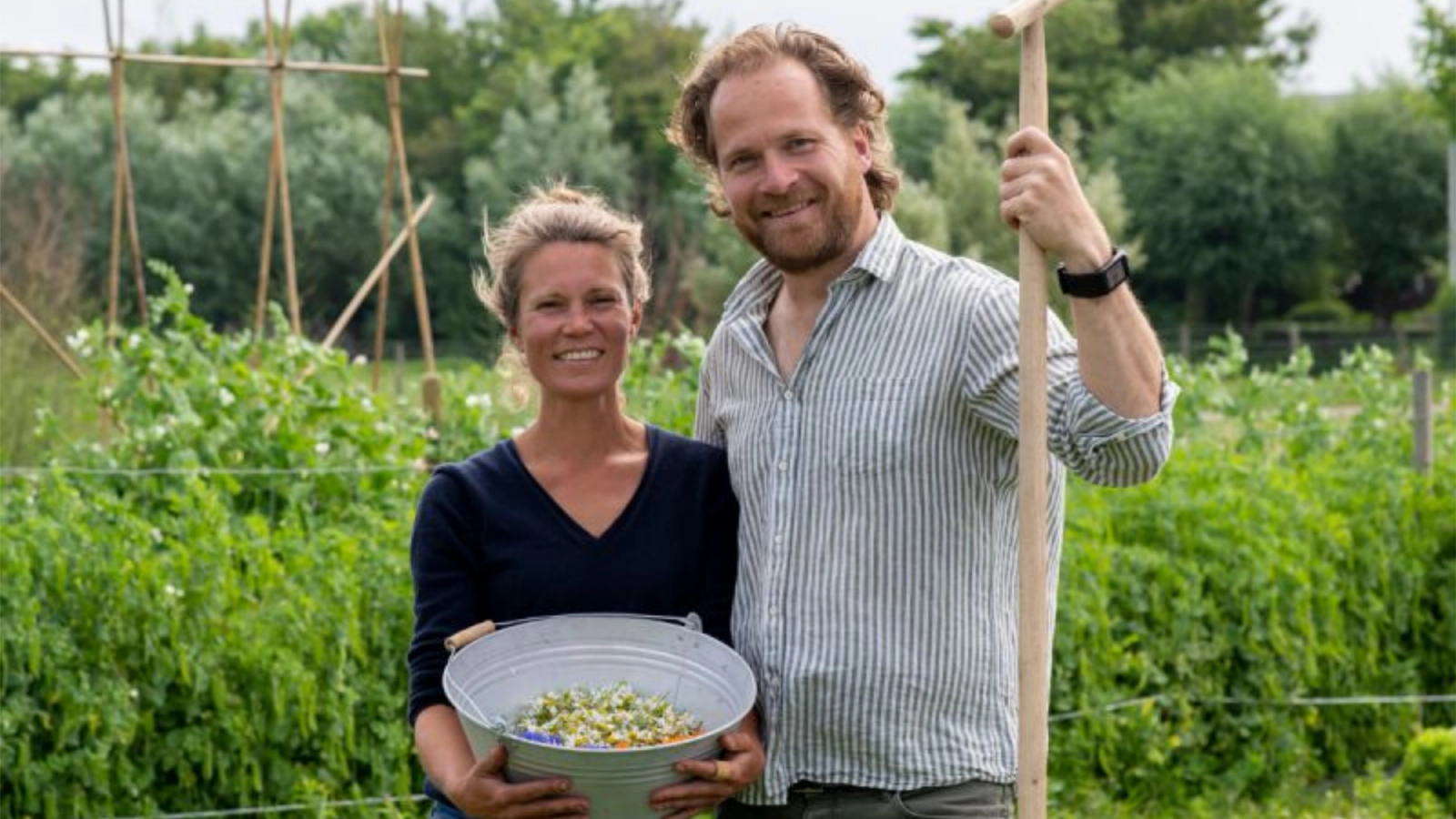 A smiling man and woman standing in a garden. The woman is holding a bucket of flowers, and the man is holding a garden tool.