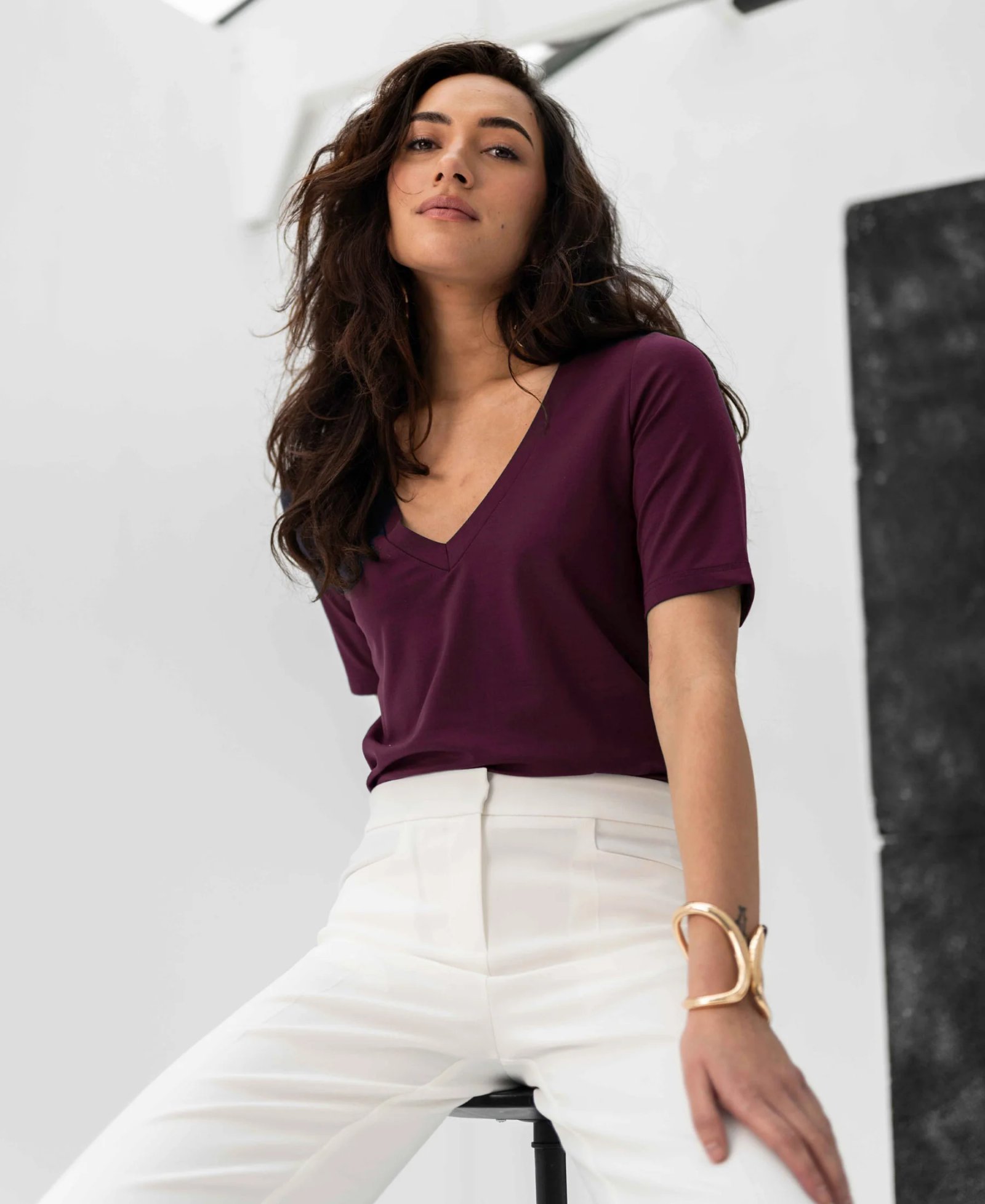A woman with long, dark, wavy hair is wearing a plum-colored V-neck T-shirt, white pants, and a gold cuff bracelet, and she is looking up.