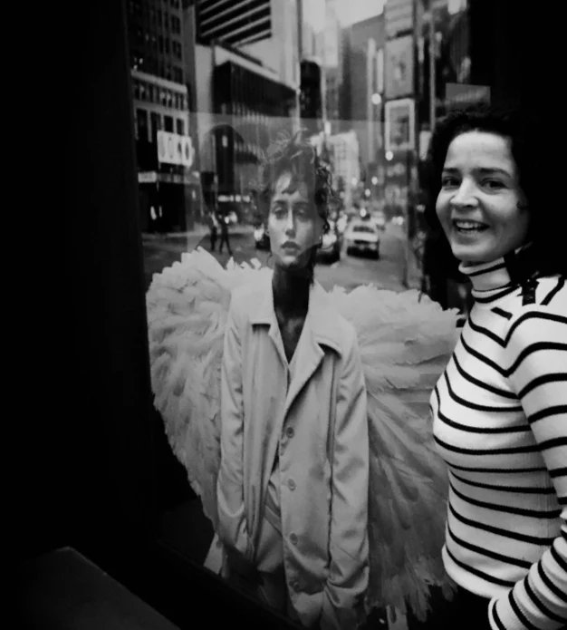 Black and white image of a smiling woman standing by a storefront window reflecting a winged figure and a city street.