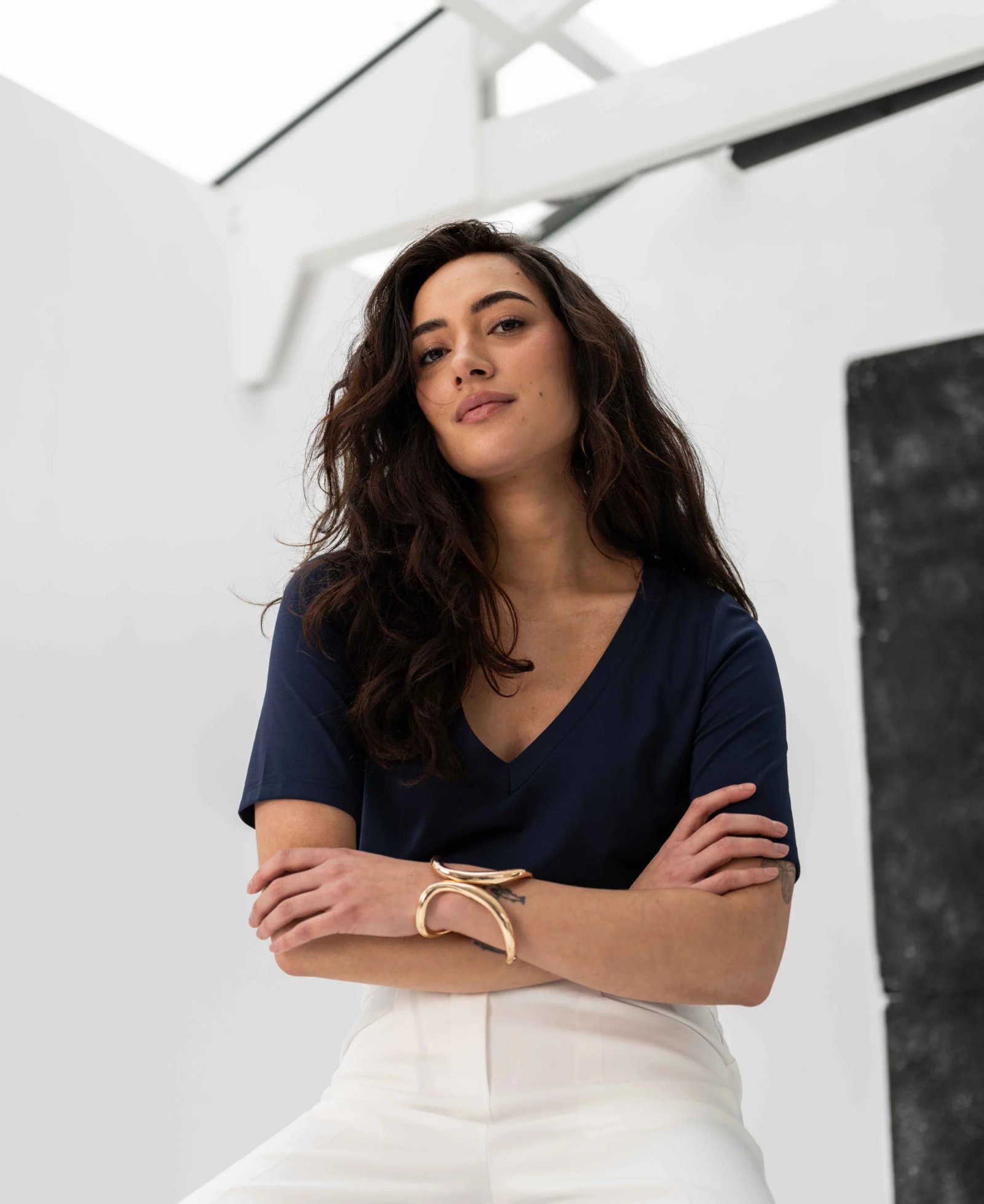 A woman with long dark hair is wearing a navy V-neck shirt, white pants, and a gold bracelet, standing with her arms crossed in a brightly lit studio.