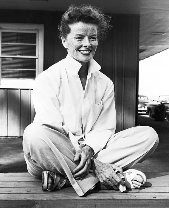 Katharine Hepburn, mit einem lächelnden, sommersprossigen Gesicht und vom Wind zerzaustem Haar, sitzt im Schneidersitz auf einer Veranda und trägt ein weißes Hemd und eine helle Hose.