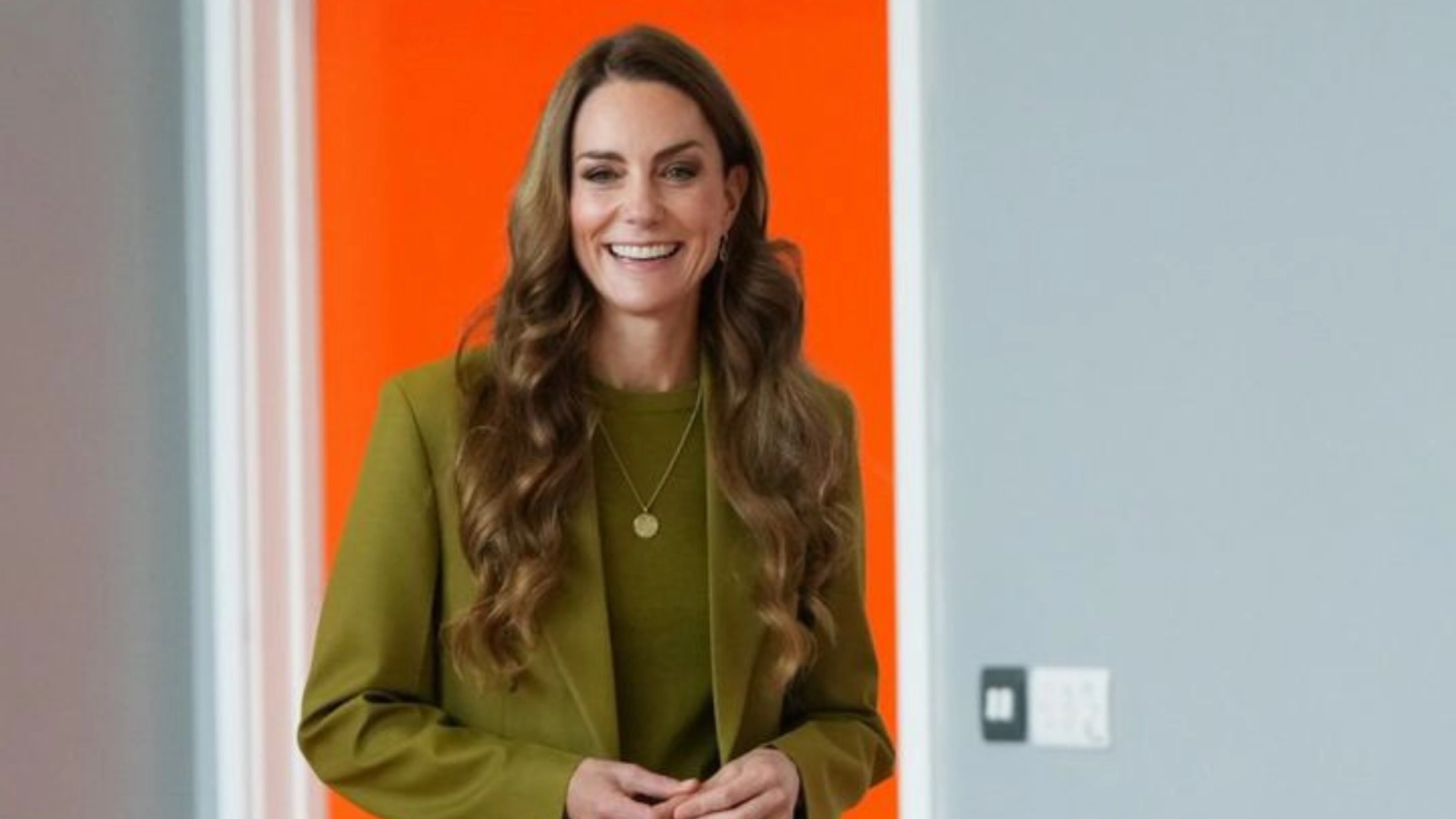 Kate Middleton lächelt in einem grünen Blazer, mit langen, welligen Haaren, vor einem orange-grauen Hintergrund.