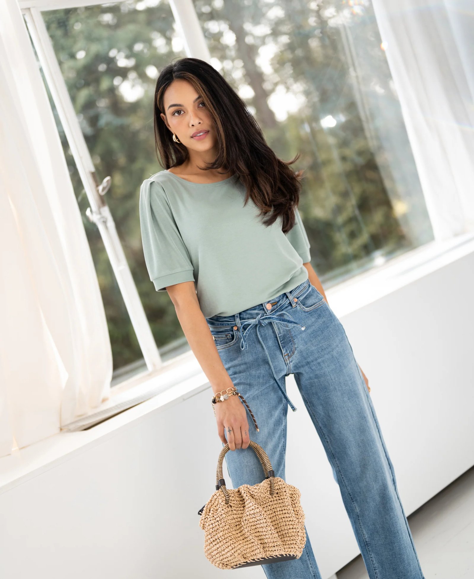 A woman wearing a light green puff-sleeve top and blue tie-waist jeans is standing by a window, holding a woven straw bag.