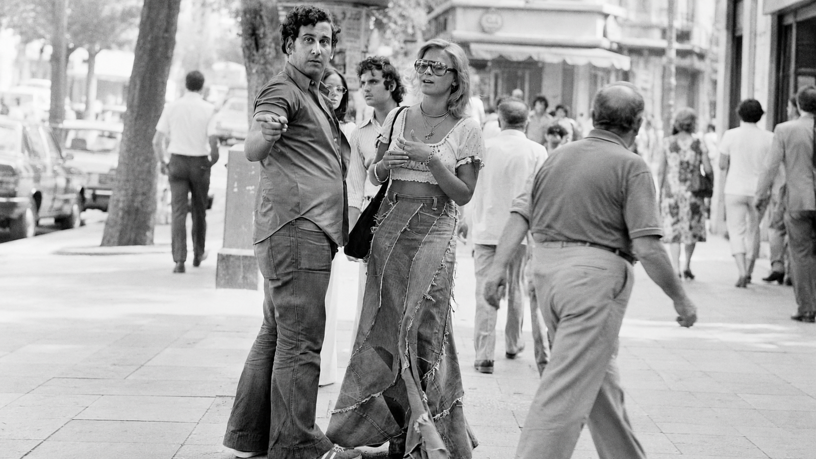 A black-and-white photo of a man pointing at the camera, standing next to a woman in a long denim skirt on a city sidewalk.