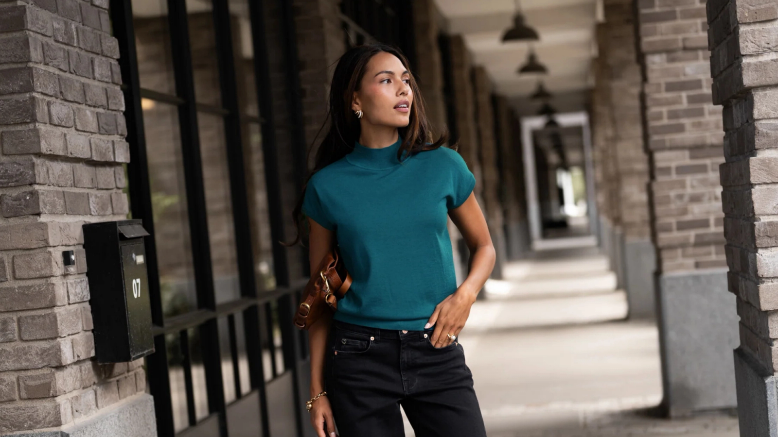 A woman wearing a teal top and black pants is standing in a brick hallway, looking to the right.