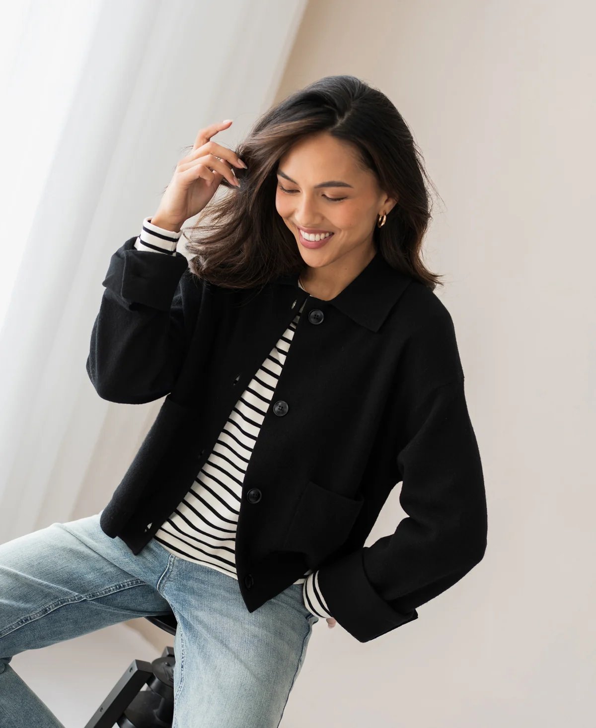Eine lächelnde Frau in einer schwarzen Jacke, einem gestreiften Hemd und Jeans streicht sich über ihr dunkles Haar.