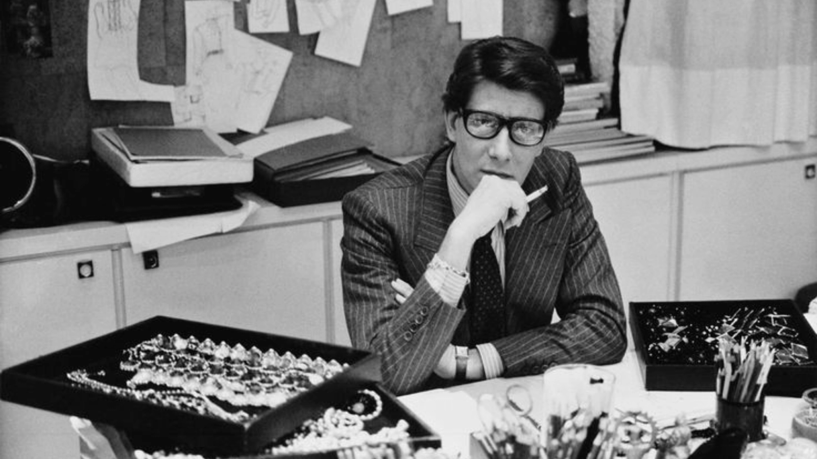 Black and white photo of Yves Saint Laurent in glasses and a pinstripe suit, holding a cigarette at a desk with jewelry trays.
