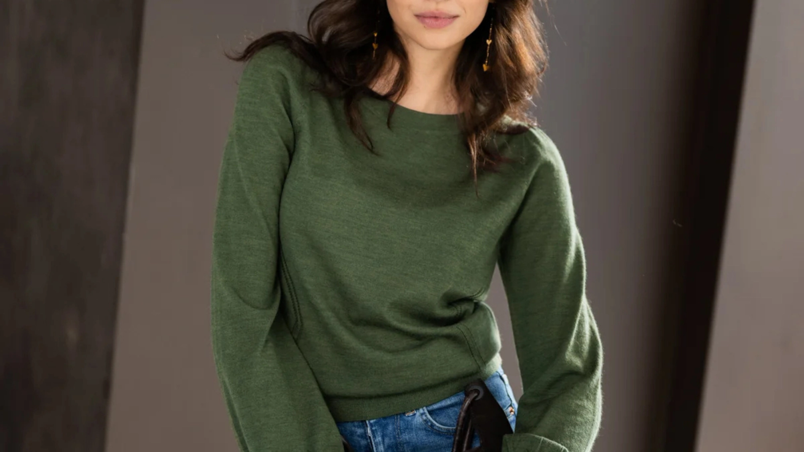 A woman wearing a dark green long-sleeve top and blue jeans, holding a black clutch.