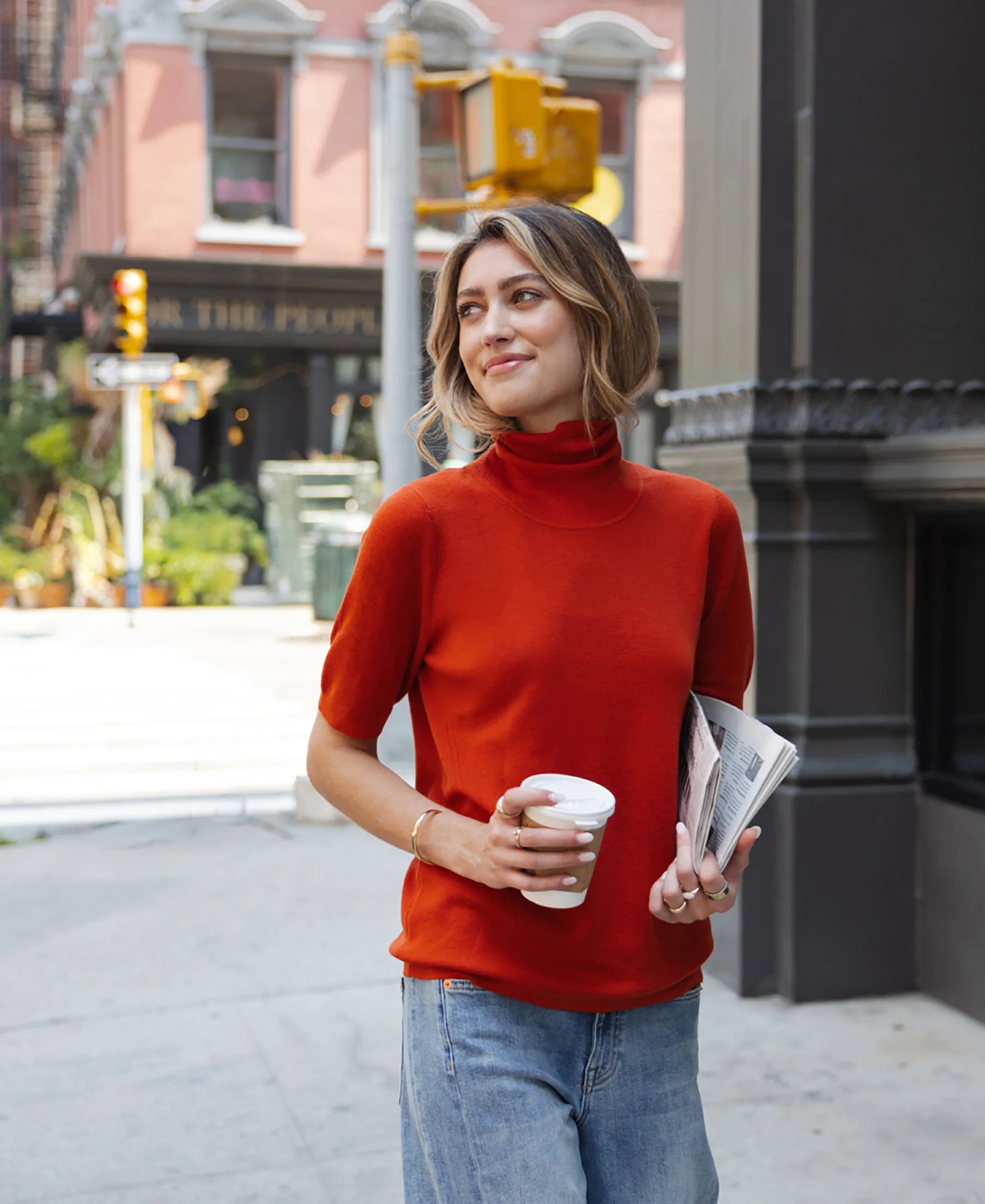 A smiling woman in an orange turtleneck is walking down a city street, holding a coffee cup and a newspaper.