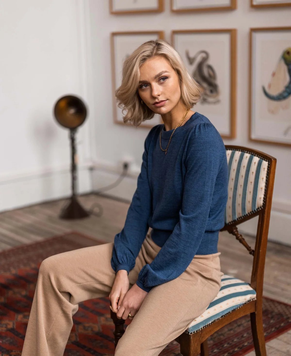A blonde woman wearing a blue top with puff sleeves and beige pants is sitting on a striped chair, glancing subtly to the side.