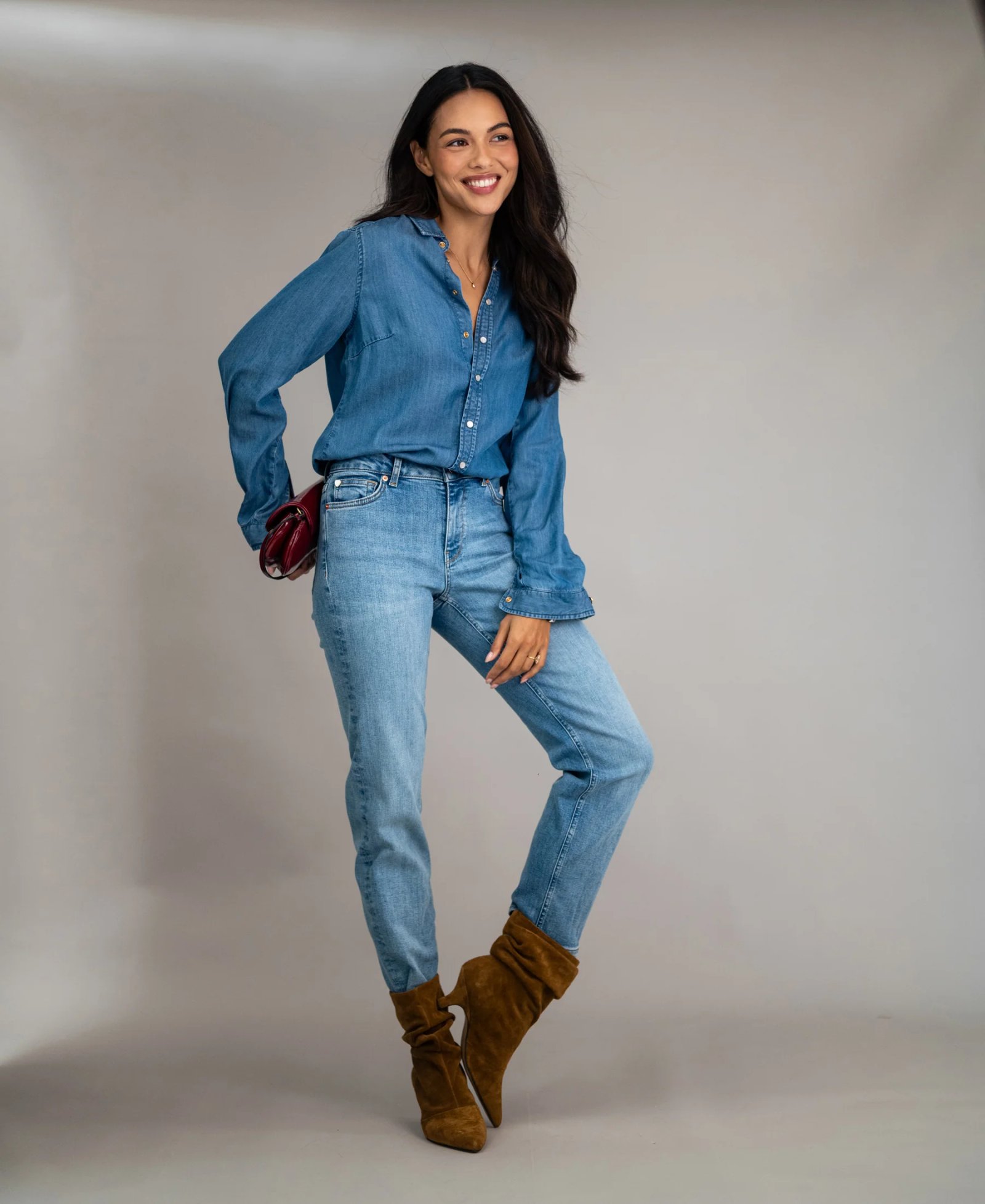 A smiling woman wearing a denim shirt, jeans, brown suede boots, and a red clutch.