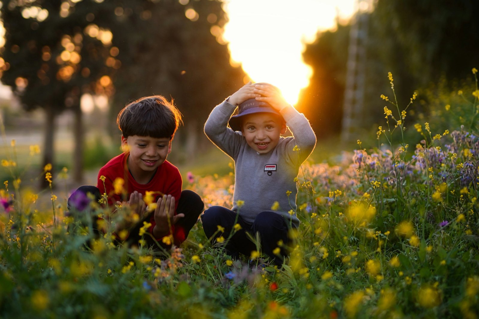 Two young boys squatting in a field of wildflowers at sunset; one looks at his hands, the other smiles widely with hands on his hat.