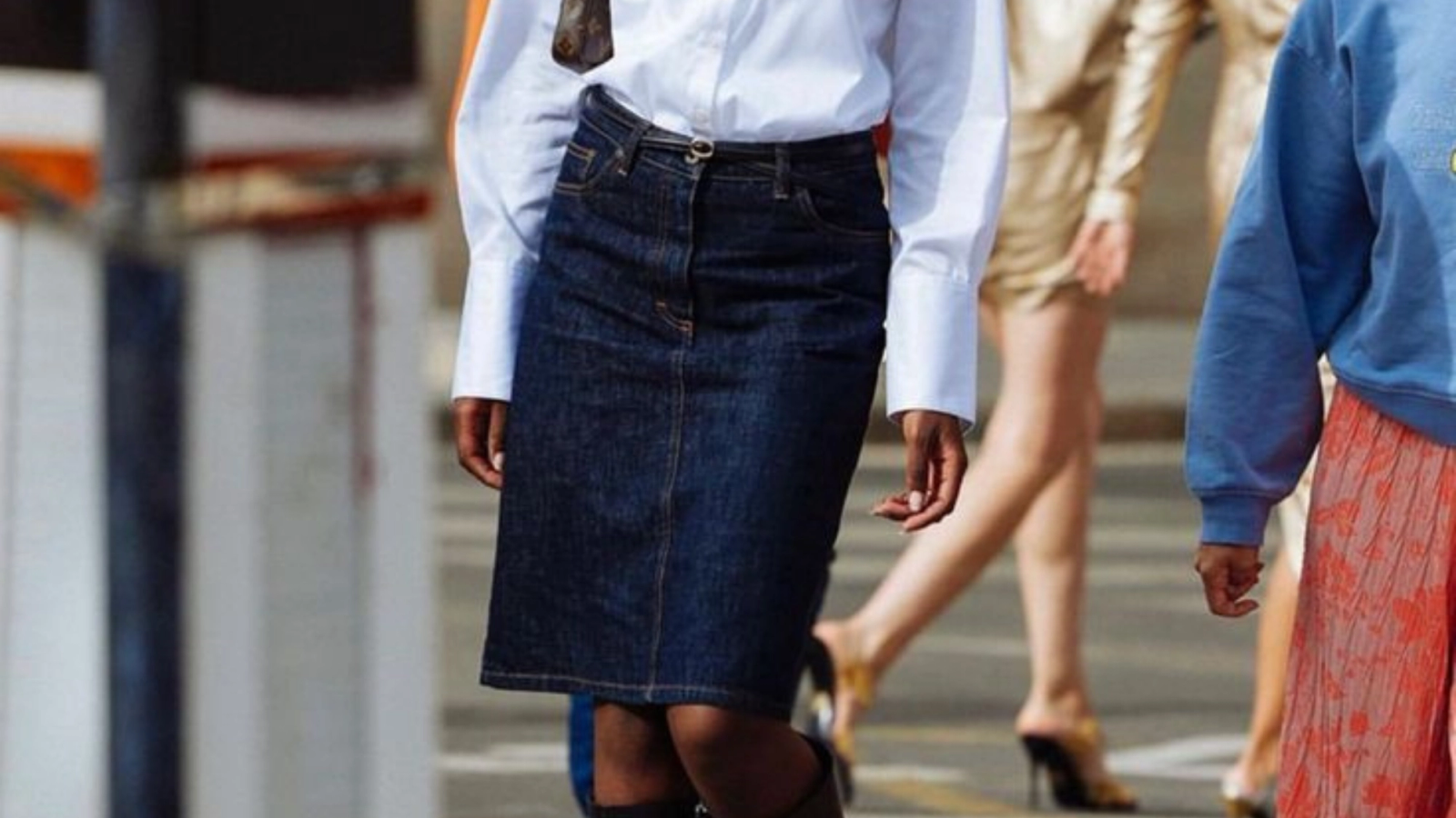 A person wearing a dark denim midi skirt and a white long-sleeved shirt is walking, with other pedestrians blurred in the background.