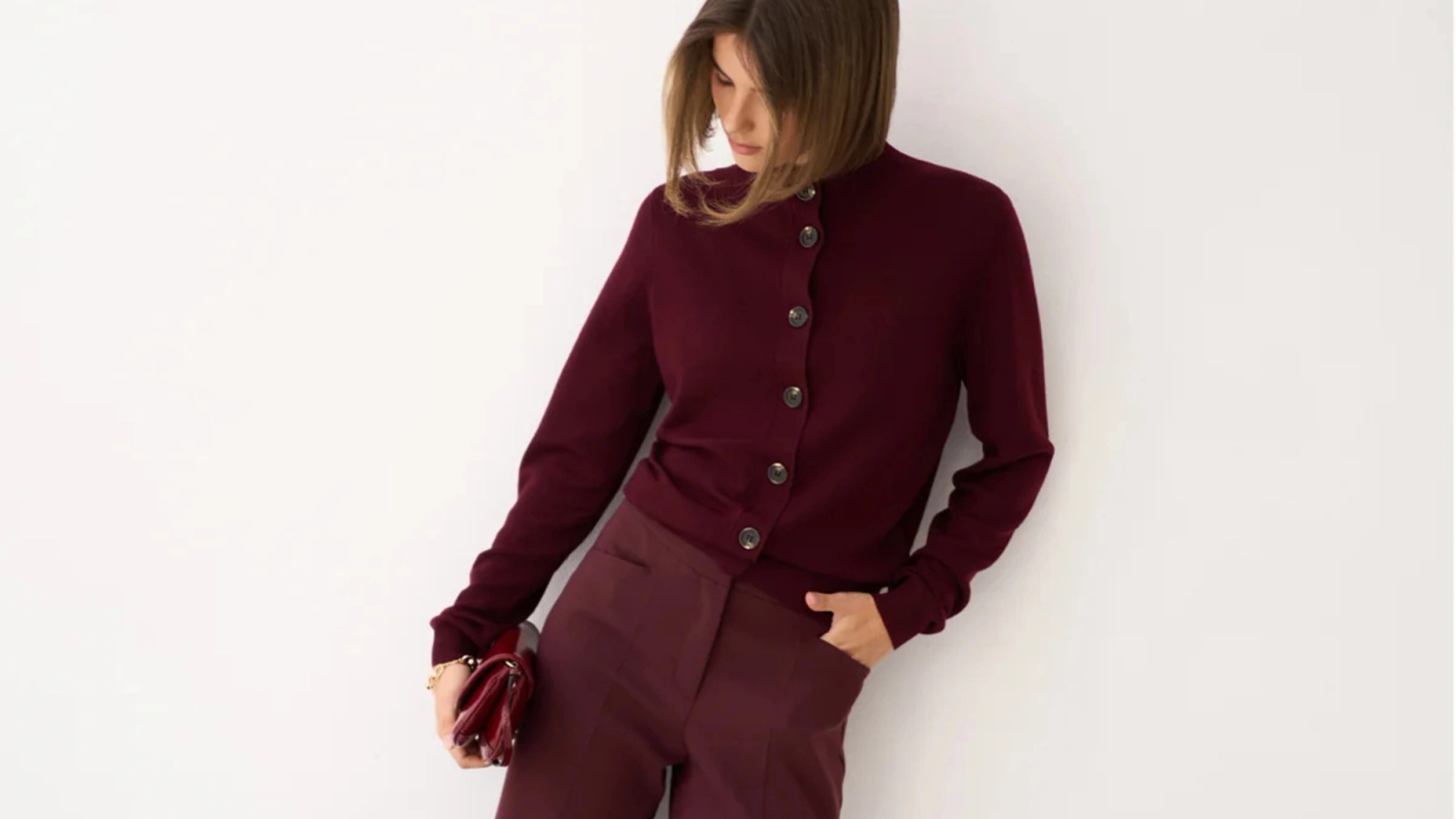 A woman in a burgundy cardigan and pants, holding a red clutch.