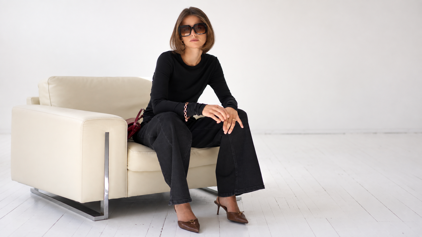 A woman in sunglasses and a black outfit sits on a cream armchair.