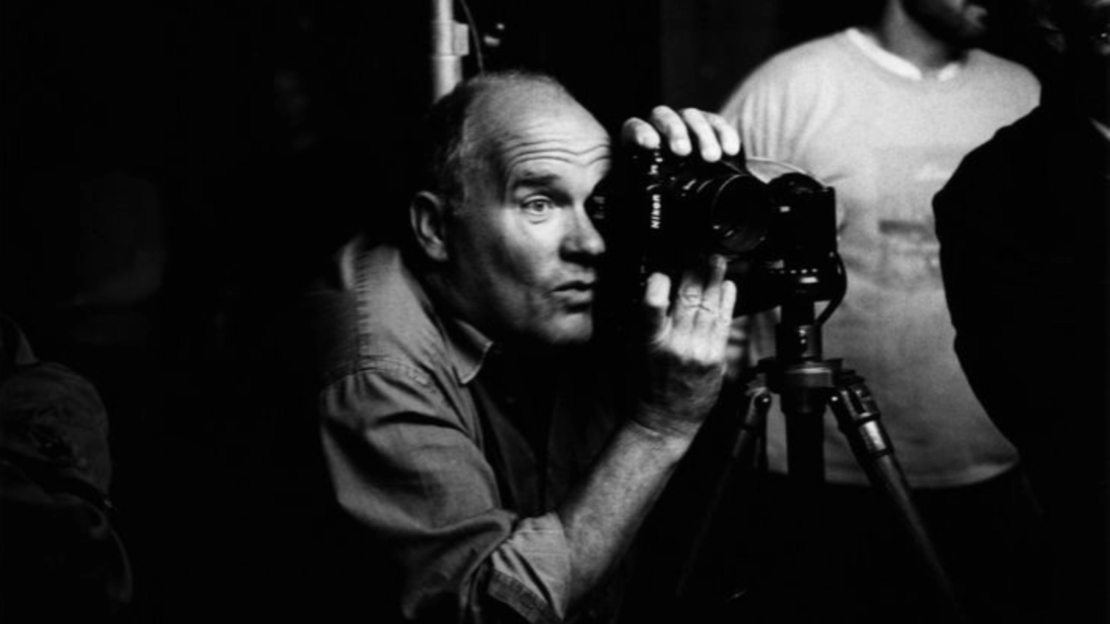 Black and white photo of a bald man looking intently through a camera mounted on a tripod.