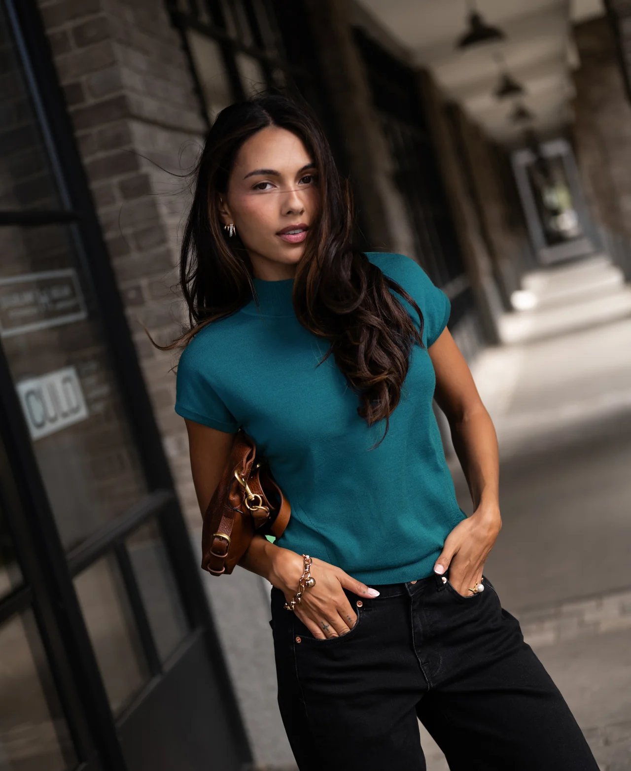 A woman in a teal mock neck top and black jeans, hands in pockets, looking at the camera in an urban setting.