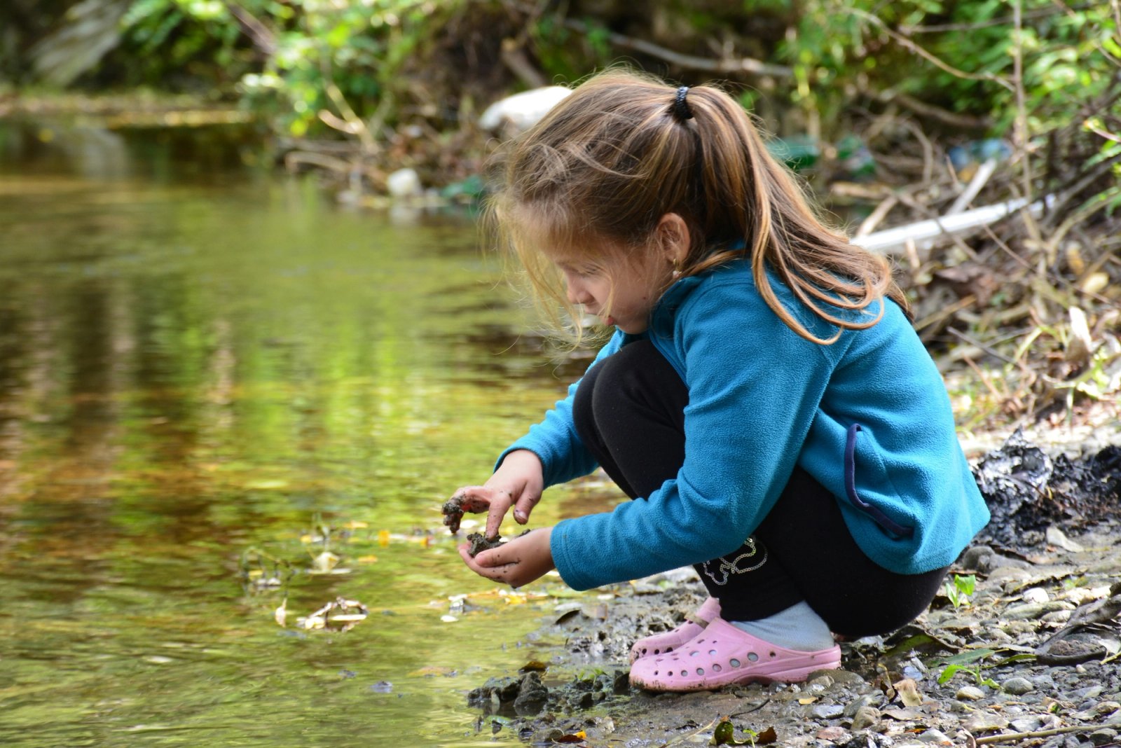 A young girl in a blue jacket crouches by a stream, intently examining dark objects in her hands.