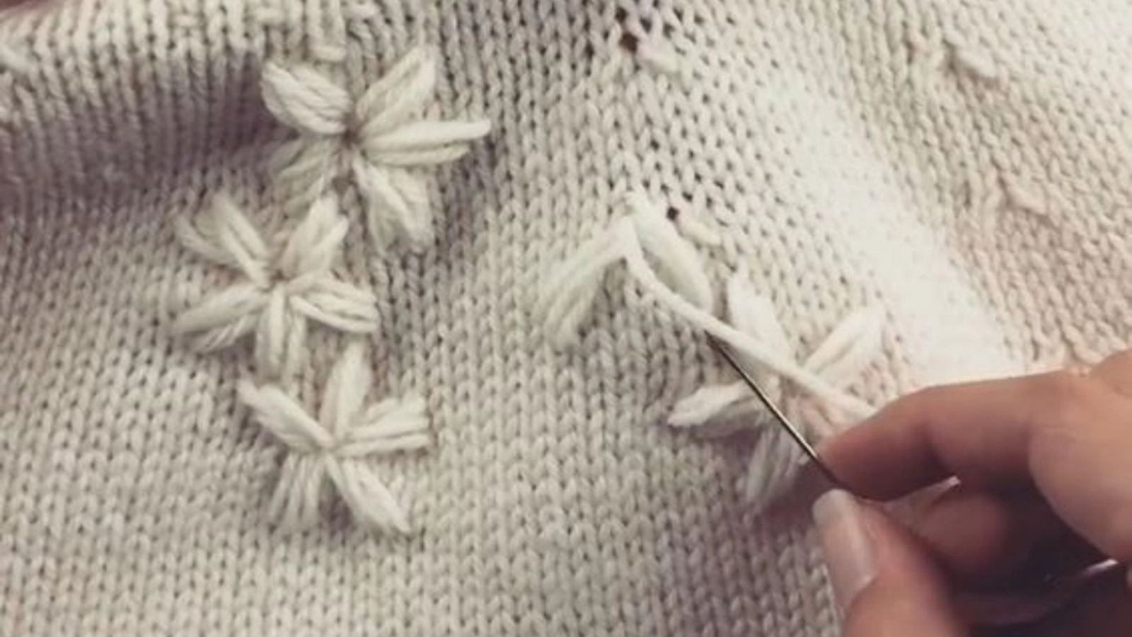 A hand is sewing white yarn flowers onto a white knitted fabric.