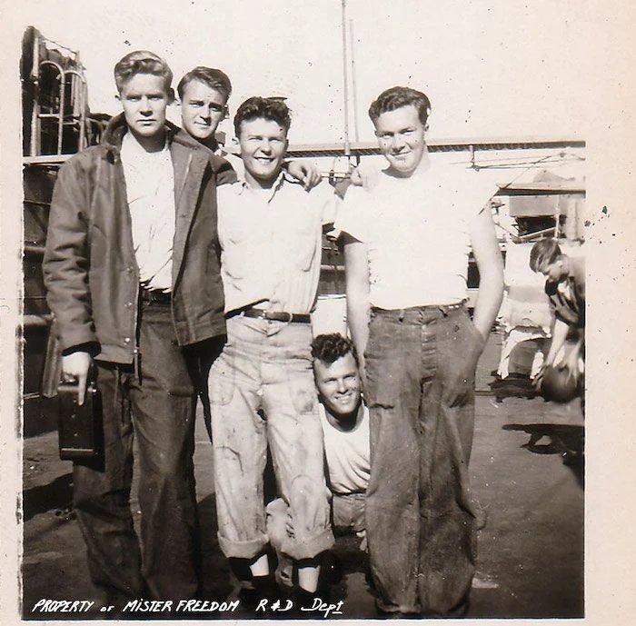 Vintage black-and-white photo of five young men posing on a deck, one of whom is crouching. Text at the bottom: 