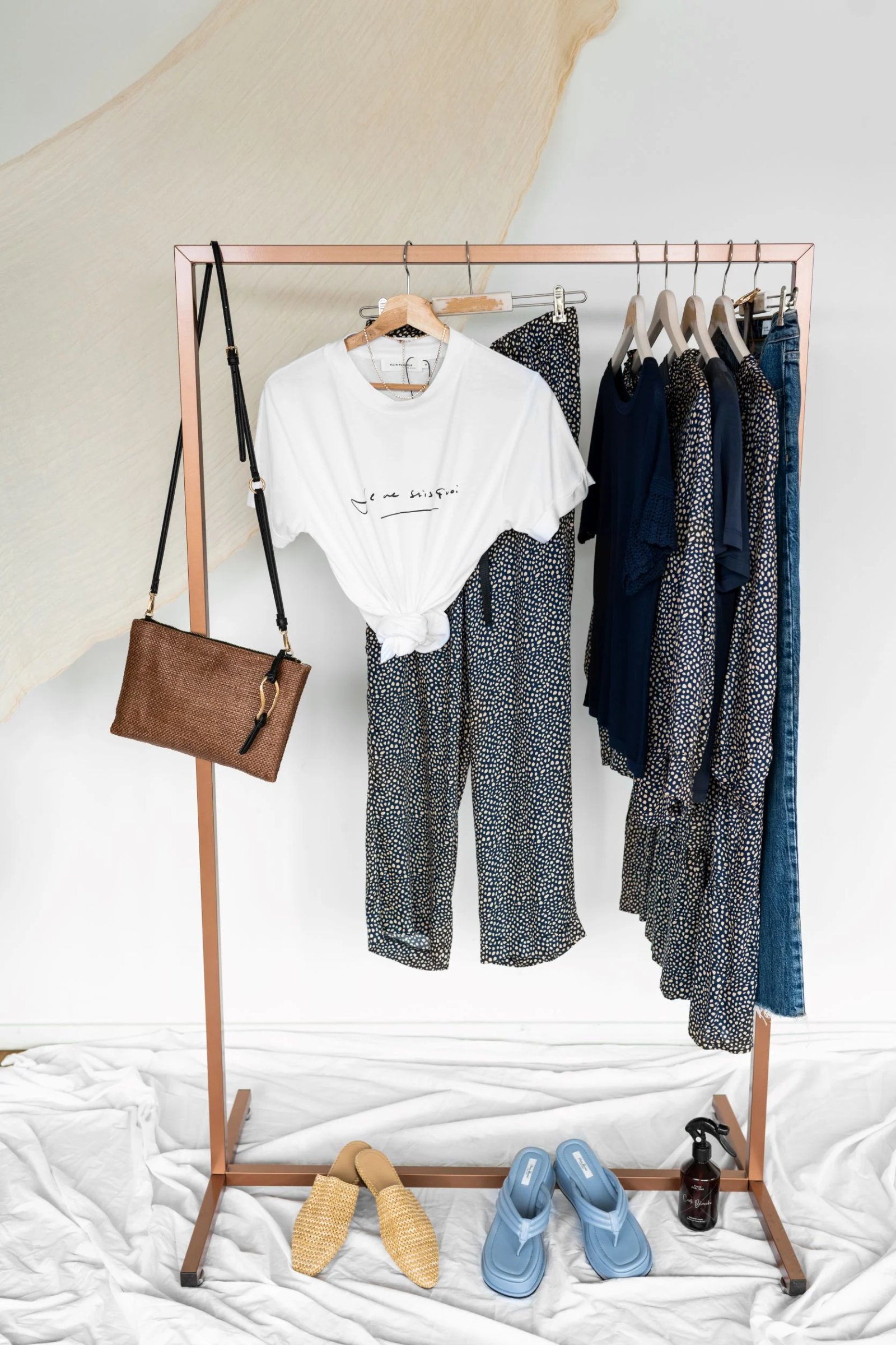 Auf einem goldfarbenen Kleiderständer hängen ein weißes T-Shirt, eine gesprenkelte Hose, verschiedene dunkle Kleidungsstücke und eine gewebte Tasche, während auf dem Boden daneben hellbraune Pantoletten und blaue Flip-Flops liegen.