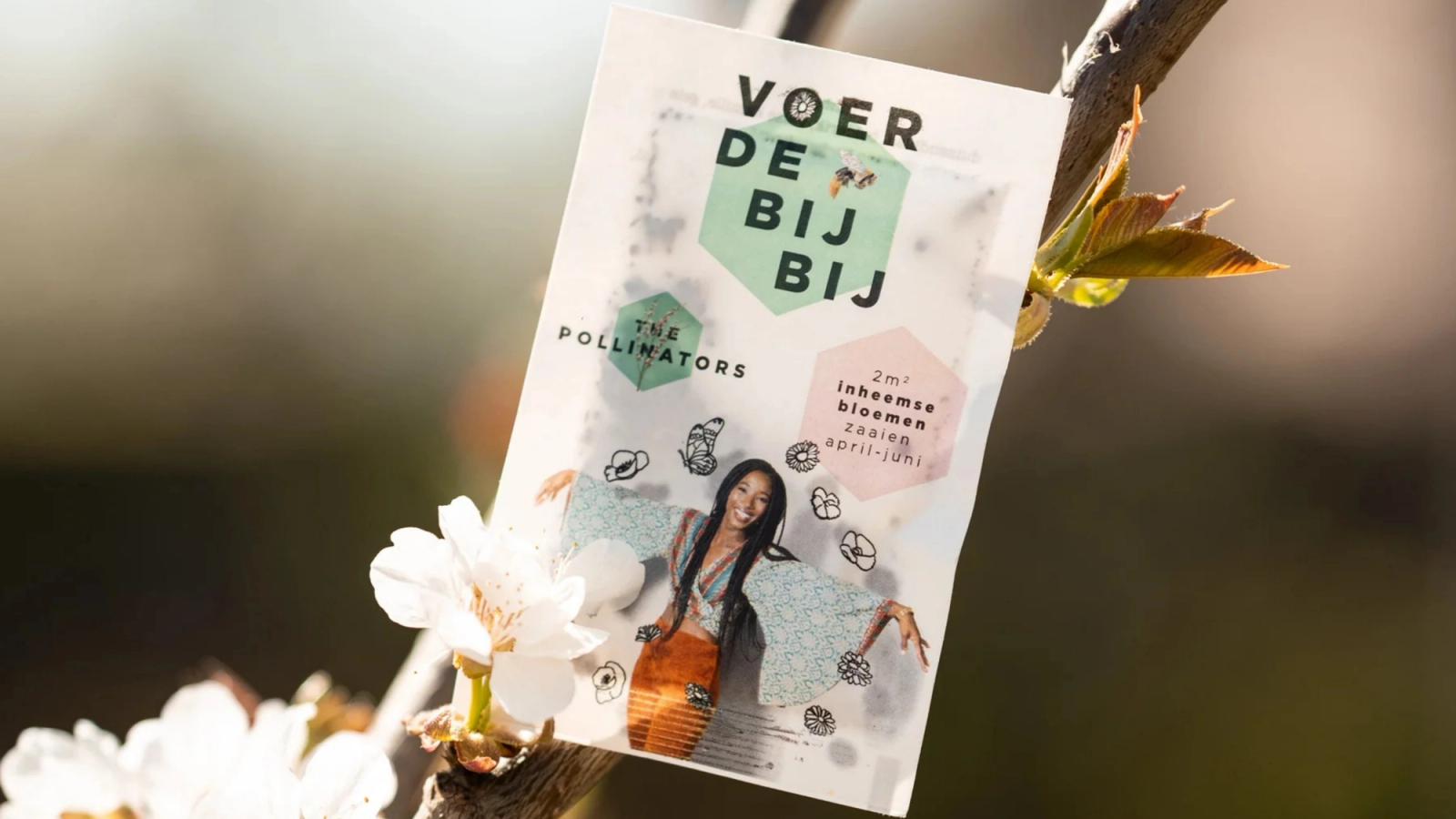 An einem blühenden Ast hängt ein Samentütchen für „The Pollinators“ mit der Aufschrift „Voer De Bij Bij“.