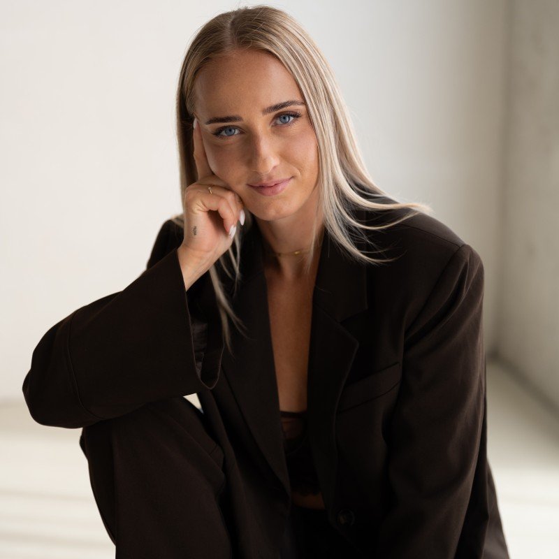 Blonde woman with blue eyes in a dark brown blazer, hand on chin, looking at the camera.