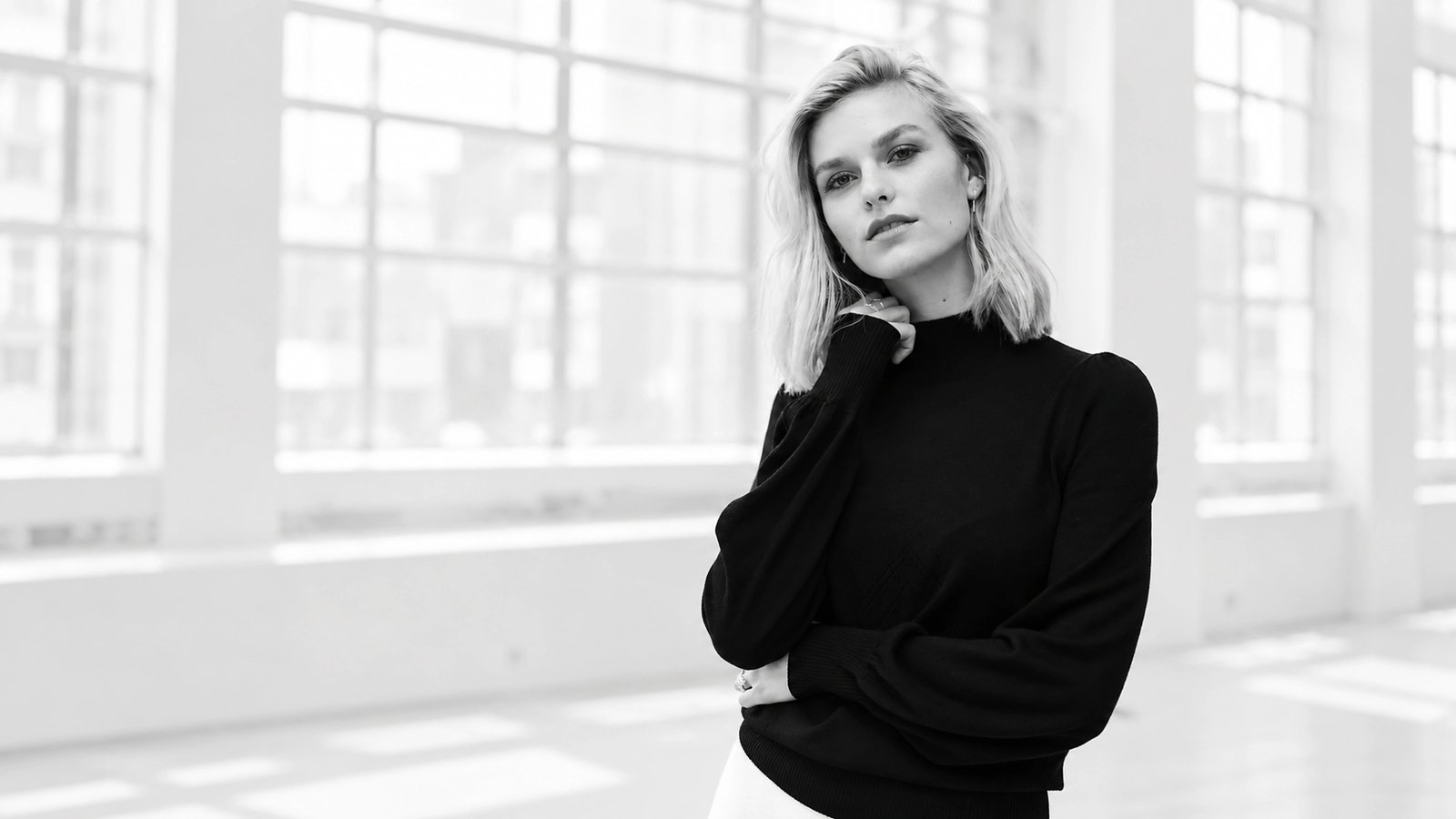 A black-and-white image of a woman with light hair wearing a dark sweater, standing in a bright room with large windows.