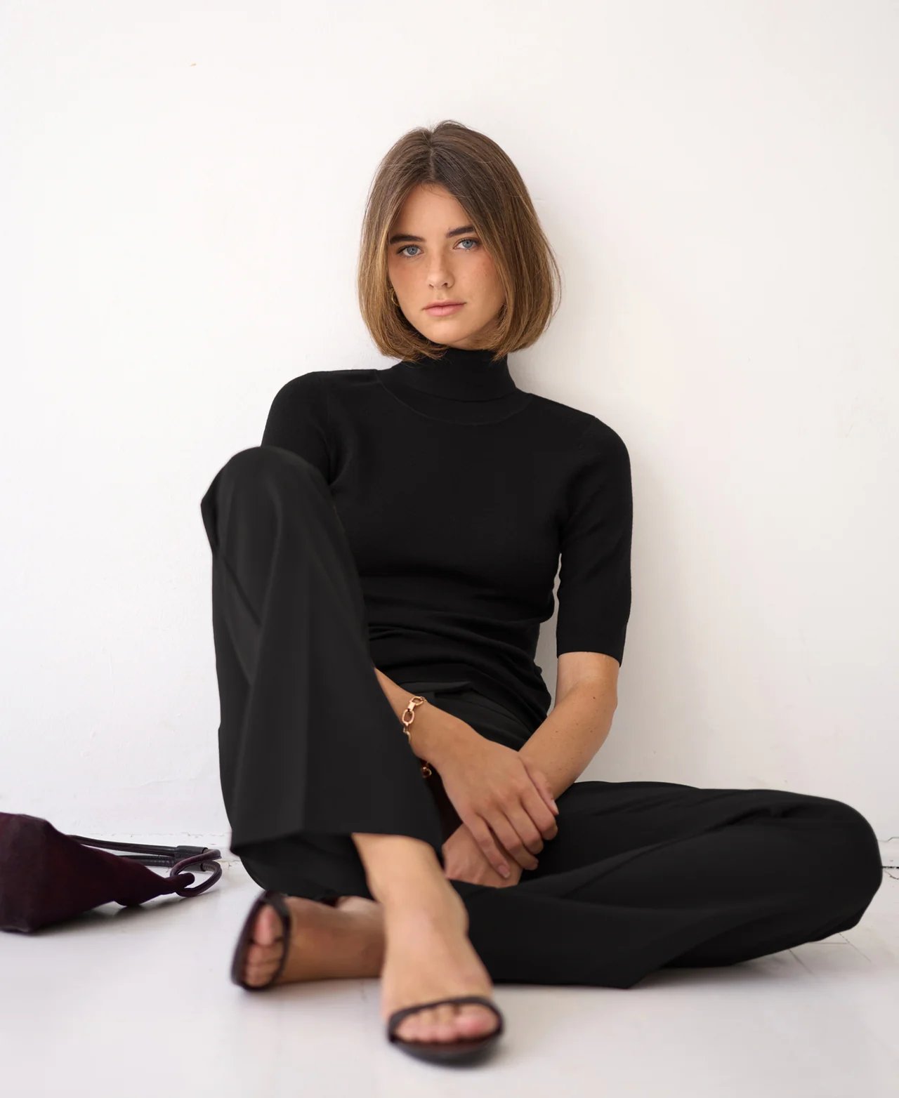 A woman with a bob haircut sits on a white floor, wearing a black turtleneck, black pants, and black sandals.