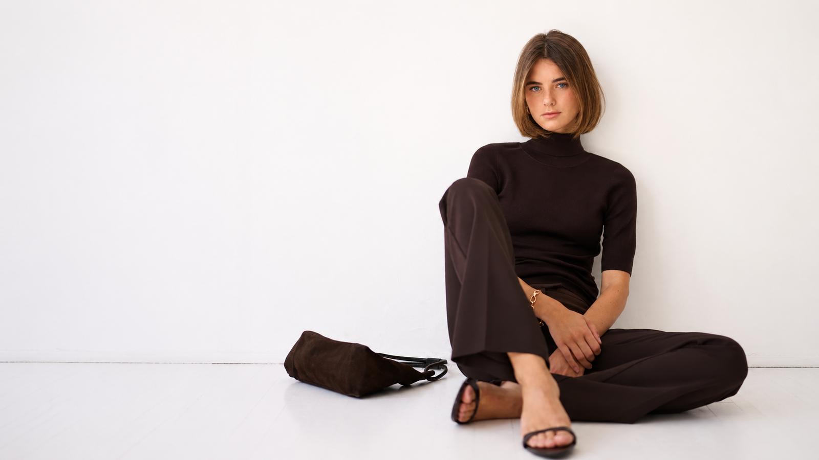 A young woman wearing a dark brown turtleneck and pants is sitting on a white floor, with a brown bag next to her.