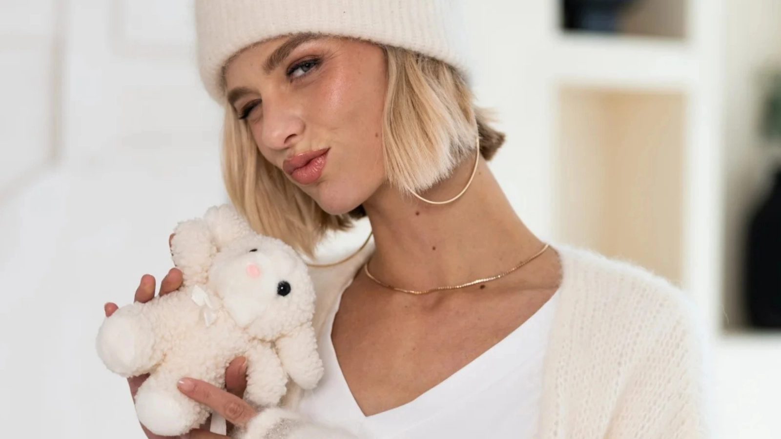 A woman wearing a white beanie and a cardigan winks and holds a white stuffed lamb.