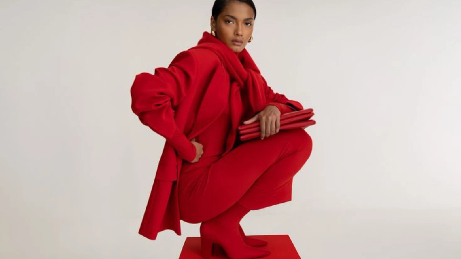 Eine Frau in einem komplett roten Outfit, bestehend aus einem voluminösen Mantel, einem Schal, einer Hose und Stiefeln, hockt auf einem roten Block und hält dabei eine rote Clutch in der Hand.