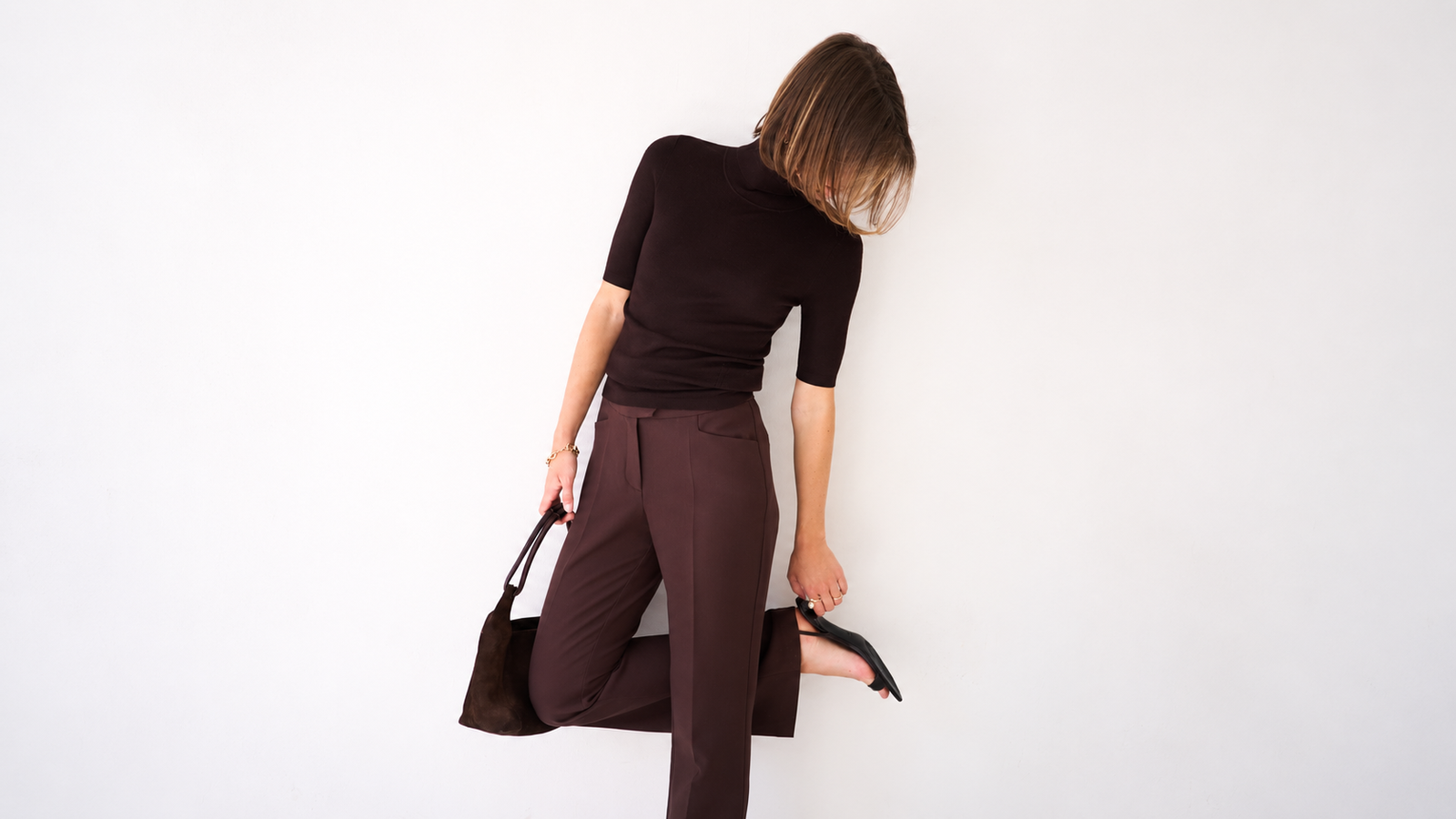 Eine Frau in einem braunen Oberteil und einer braunen Hose, die eine braune Tasche in der Hand hält und einen Fuß mit einem schwarzen Schuh anhebt.